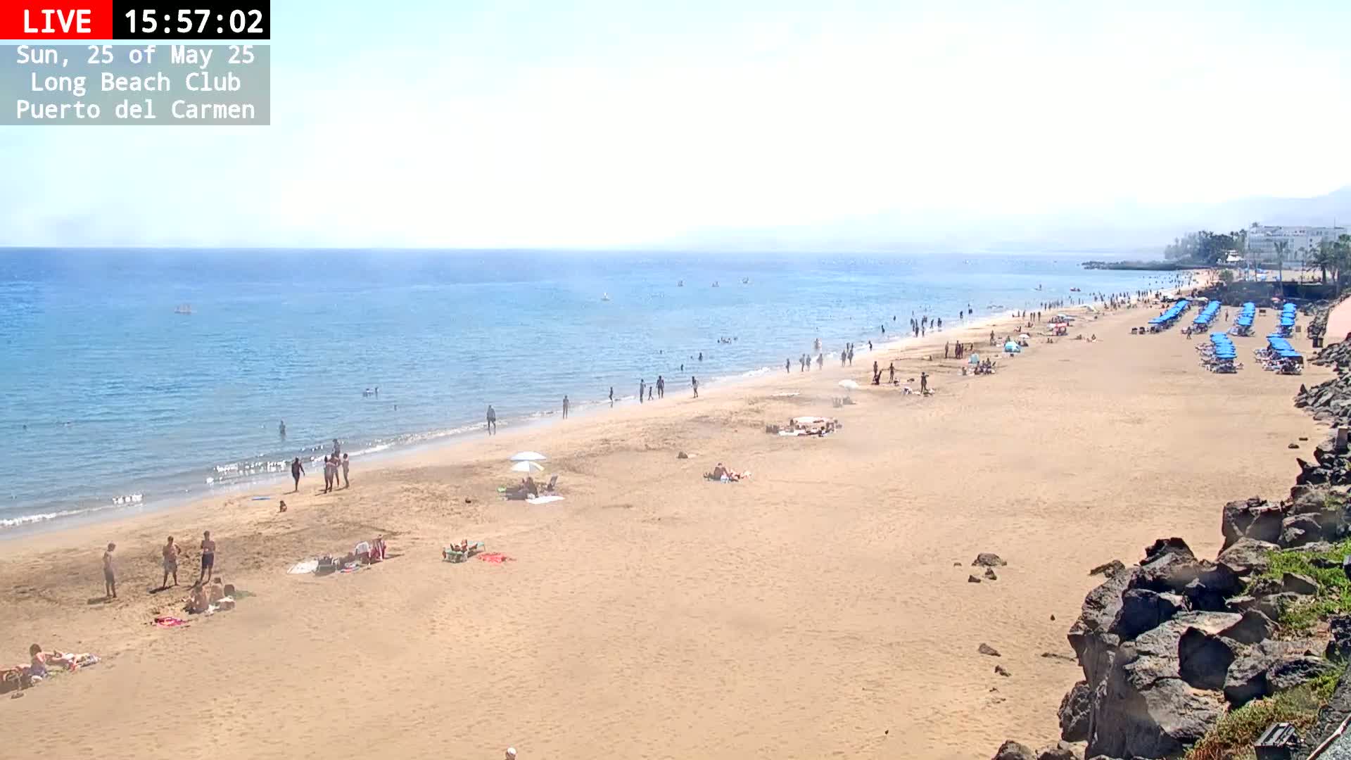 A sandy beach with numerous people sunbathing and swimming in a calm, clear ocean under a bright, sunny sky.