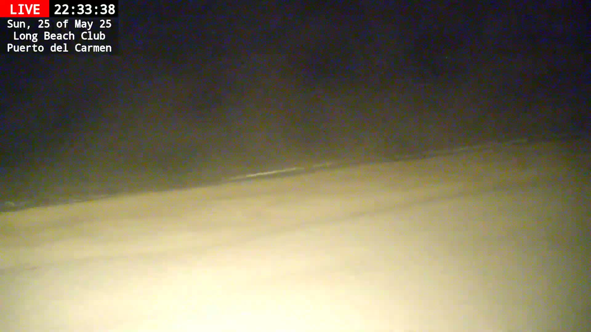A dark, nighttime beach scene shows a sandy shoreline meeting a dark ocean under a mostly clear sky.