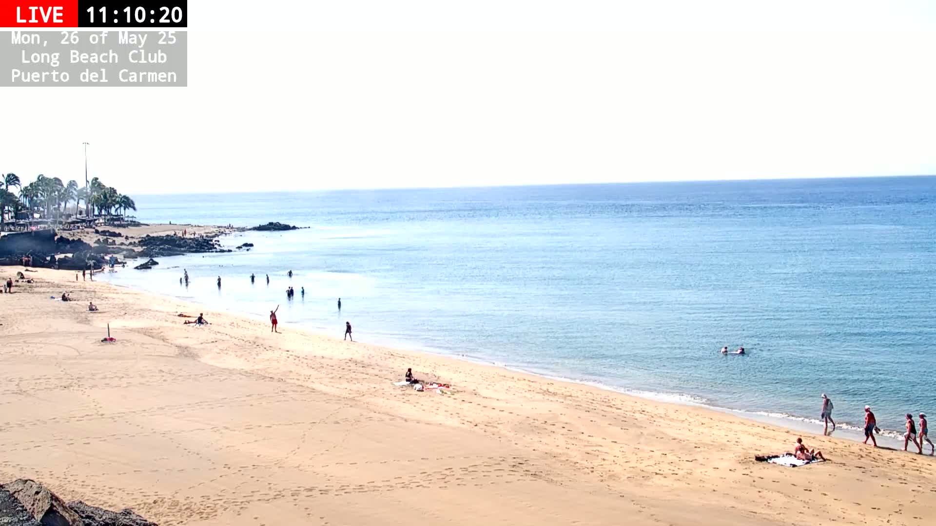 A sandy beach with calm, light-blue water under a clear sky has a few people swimming, sunbathing, and walking along the shore.