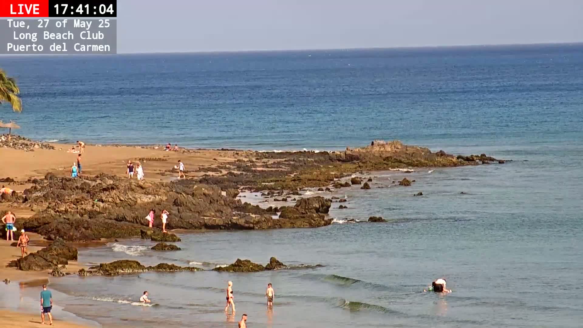 A sandy beach with dark rocks extending into a calm, light blue ocean, where several people are swimming, sunbathing, and walking.