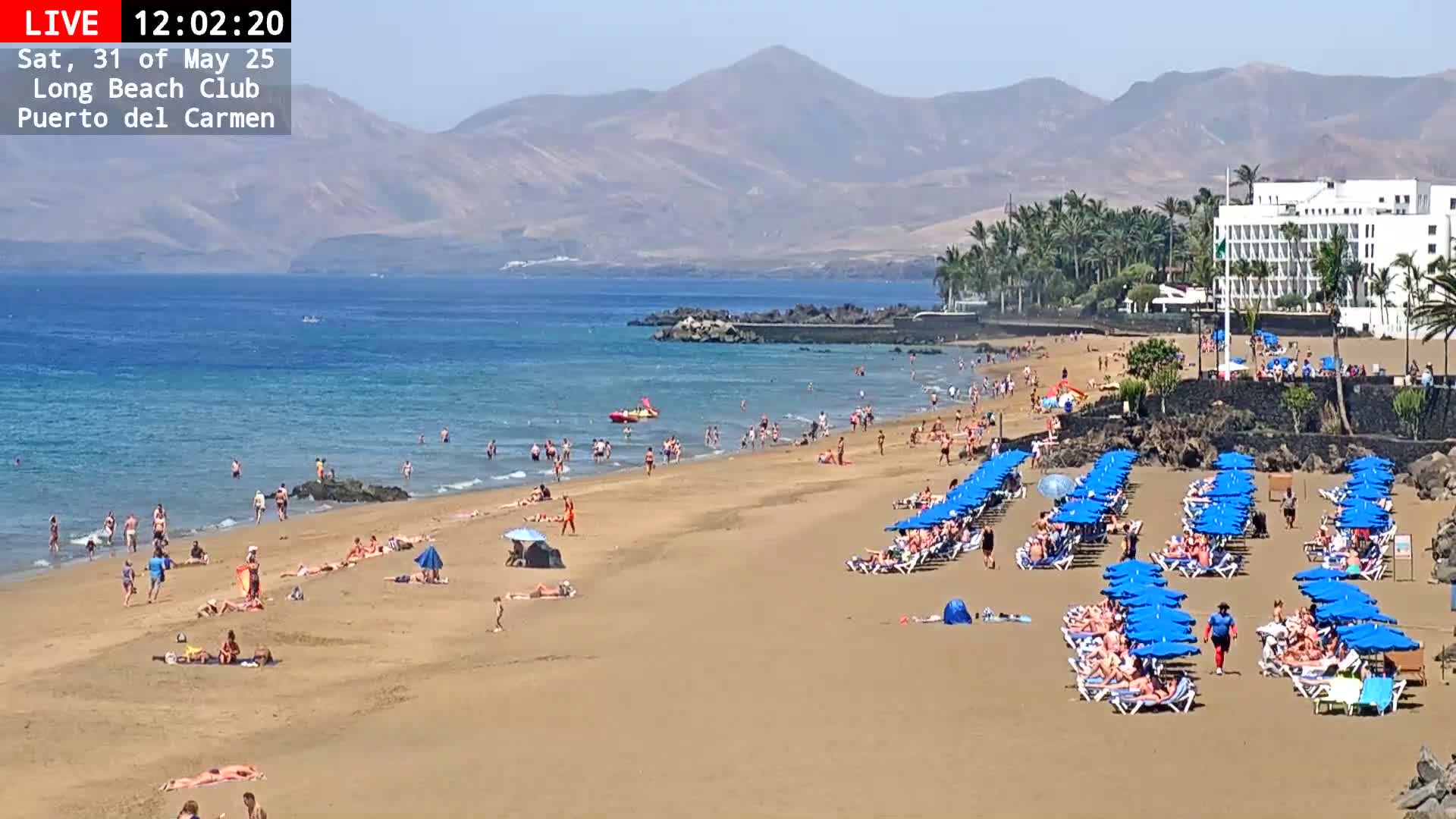 A sunny beach scene shows many people swimming, sunbathing, and lounging under blue umbrellas on a sandy beach, with calm ocean water, mountains in the background, and a hotel partially visible.