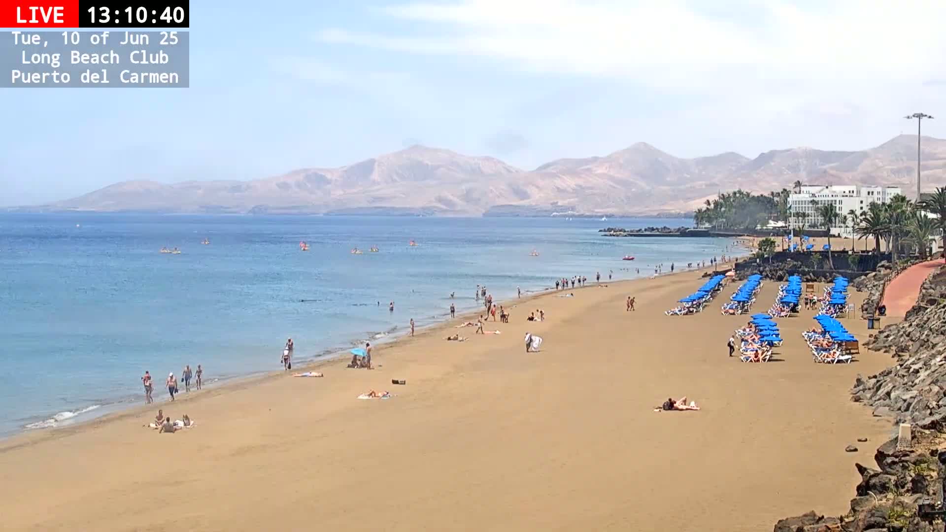 A sandy beach with numerous people sunbathing and swimming in calm, clear water, under a sunny sky, with mountains in the background.
