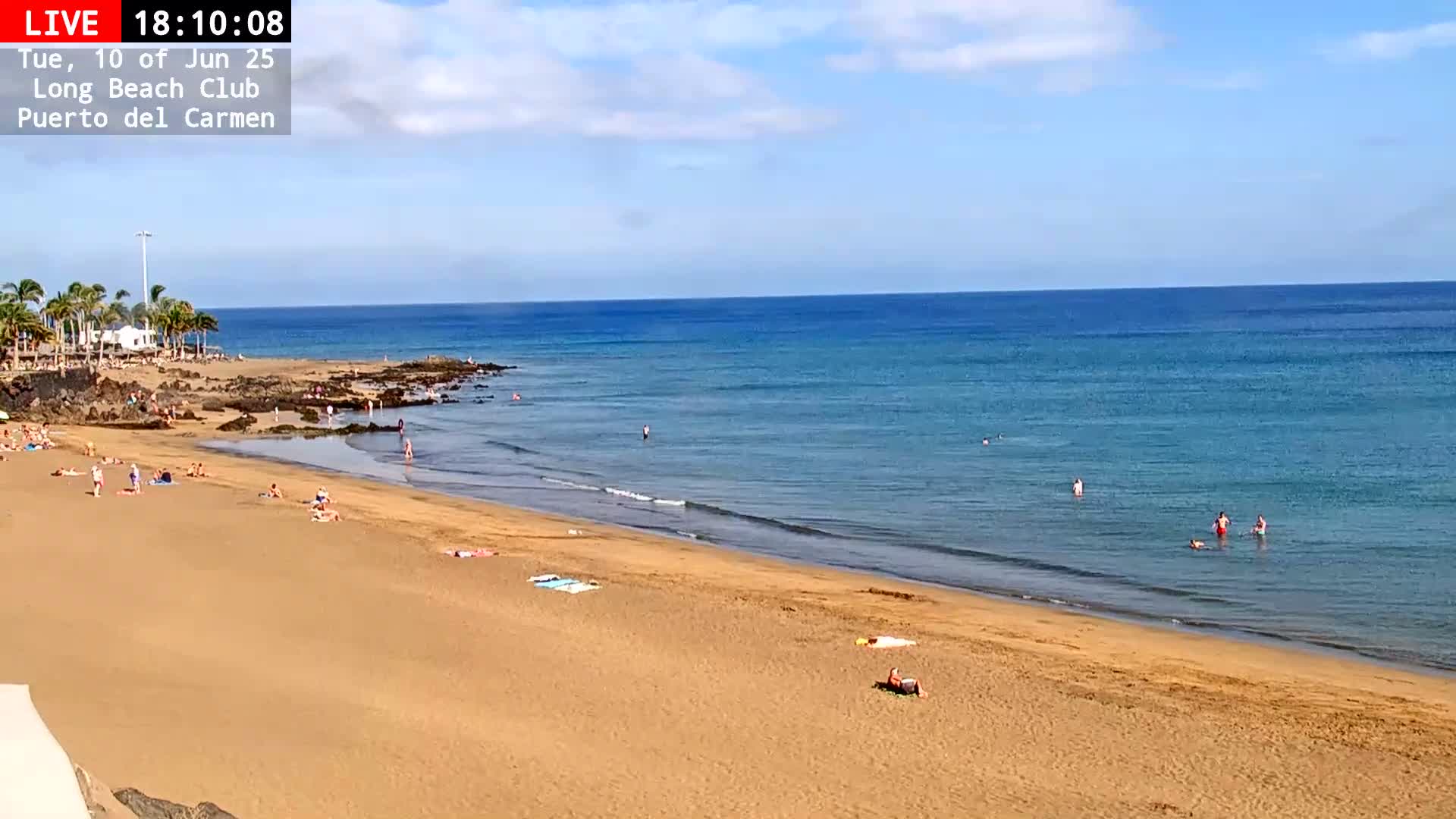 A sandy beach with scattered people sunbathing and swimming in a calm, clear blue ocean under a mostly sunny sky.