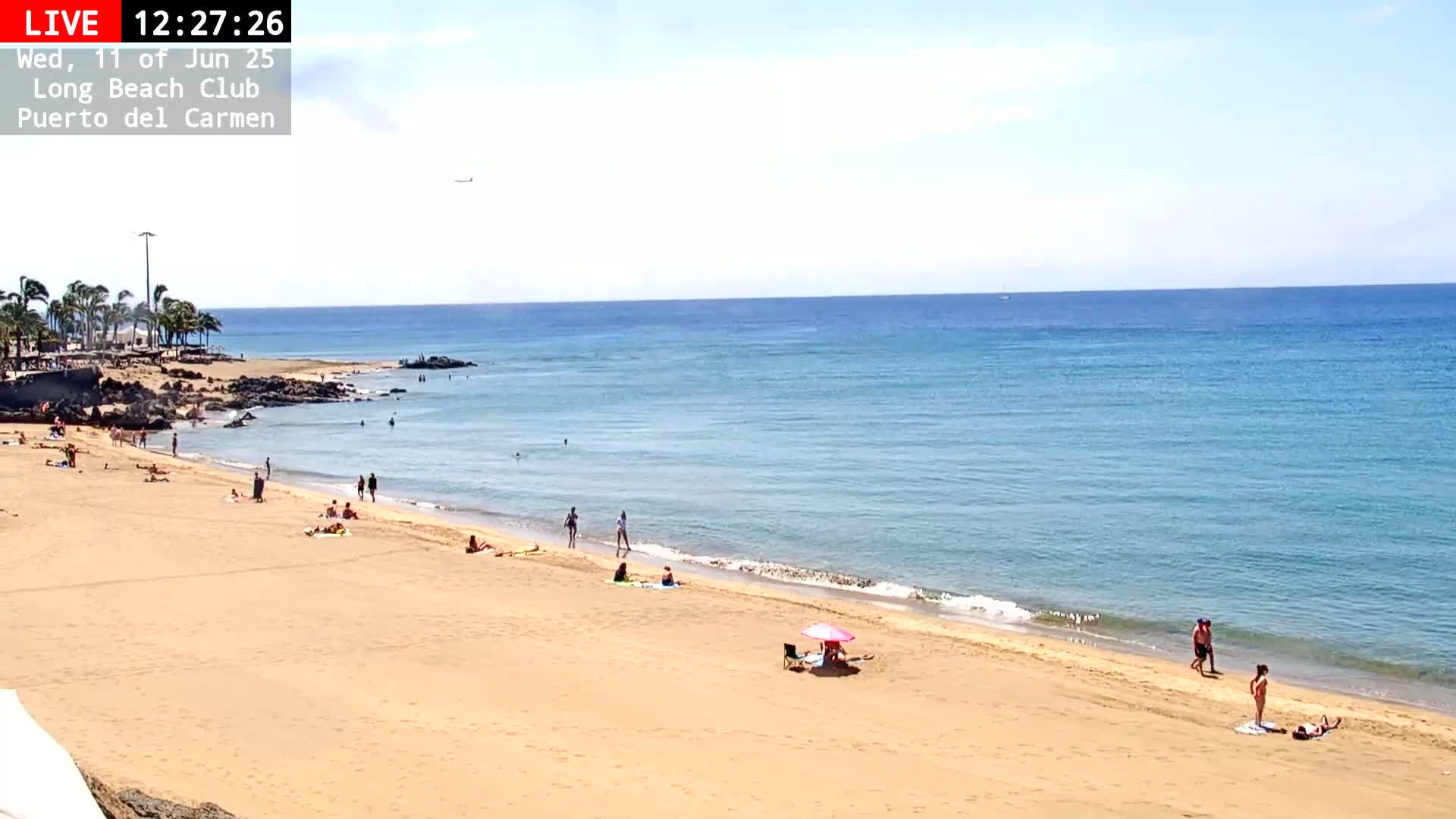 A sandy beach with scattered people is shown under a clear sky, next to a calm, blue ocean.