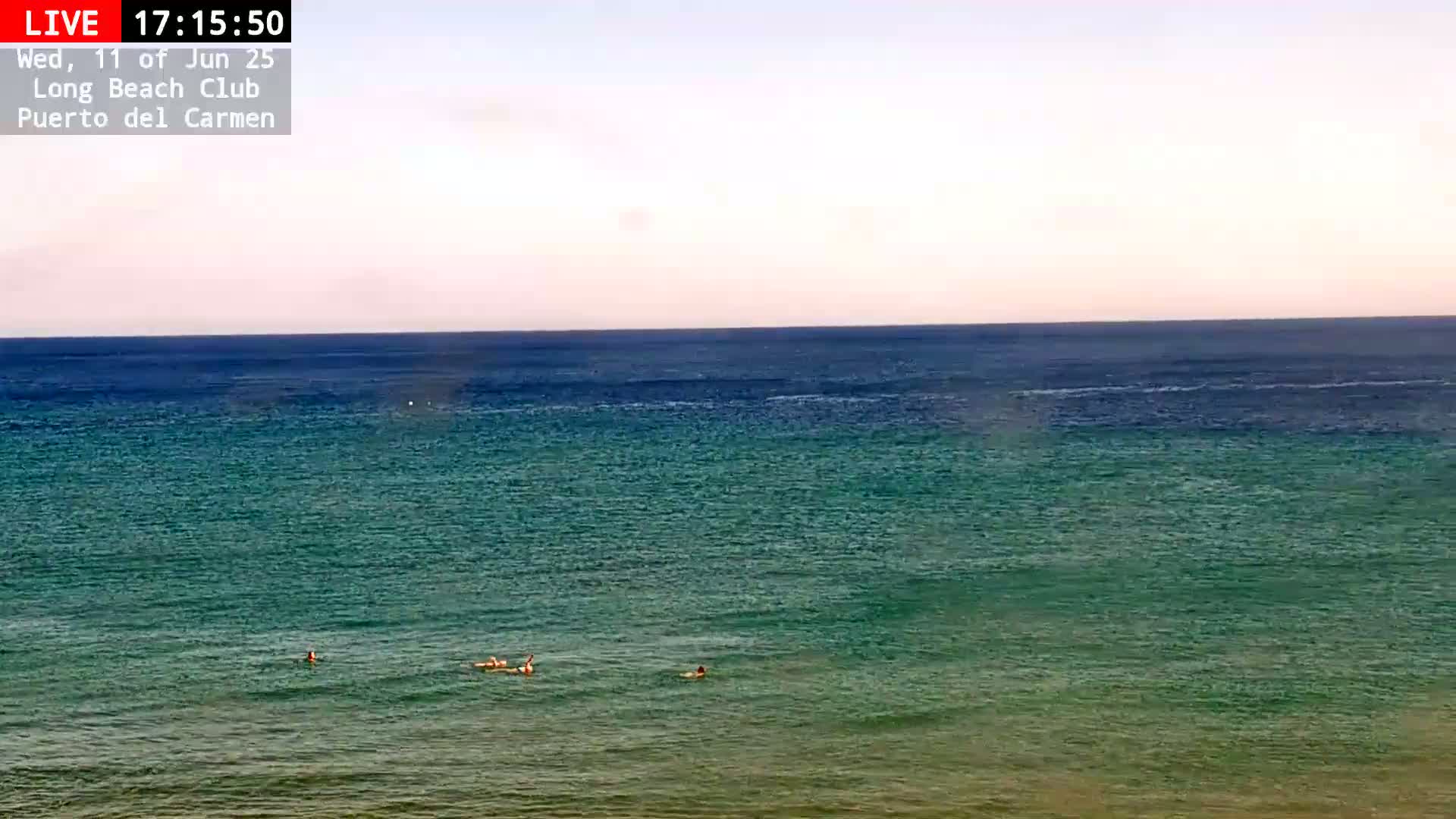 A calm, teal-green ocean under a pale sky with a few people swimming near the shore.