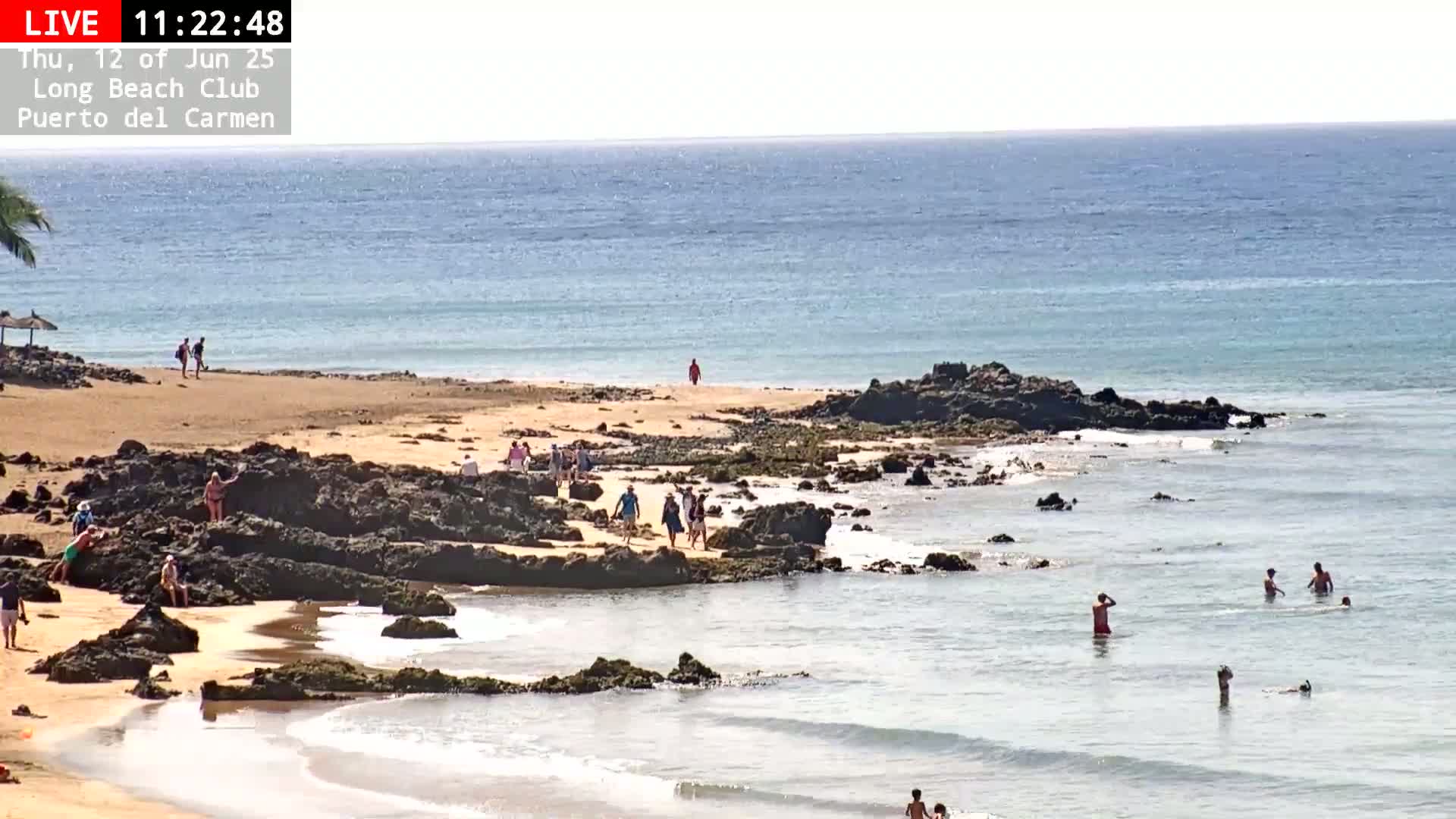 A sandy beach with dark rocks and calm, light-blue ocean water is populated by several people swimming and walking along the shore under a sunny sky.