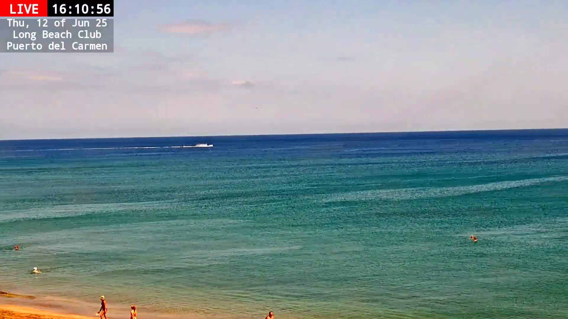A mostly sunny day shows a wide expanse of teal ocean water with a sandy beach in the foreground where several people are swimming and walking.