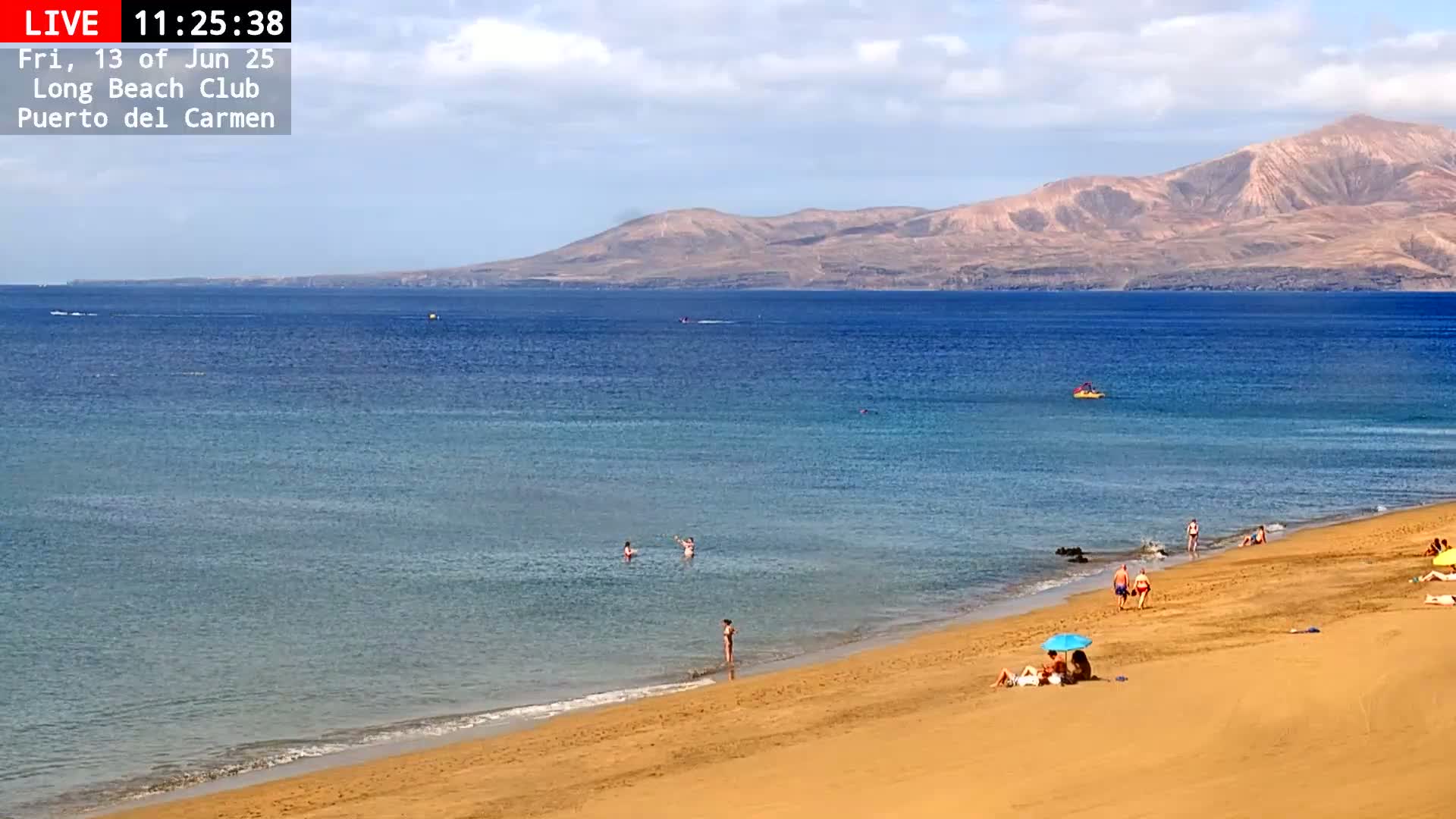 A sandy beach with a few people on it stretches to a calm, blue ocean under a partly cloudy sky, with mountains visible in the distance.