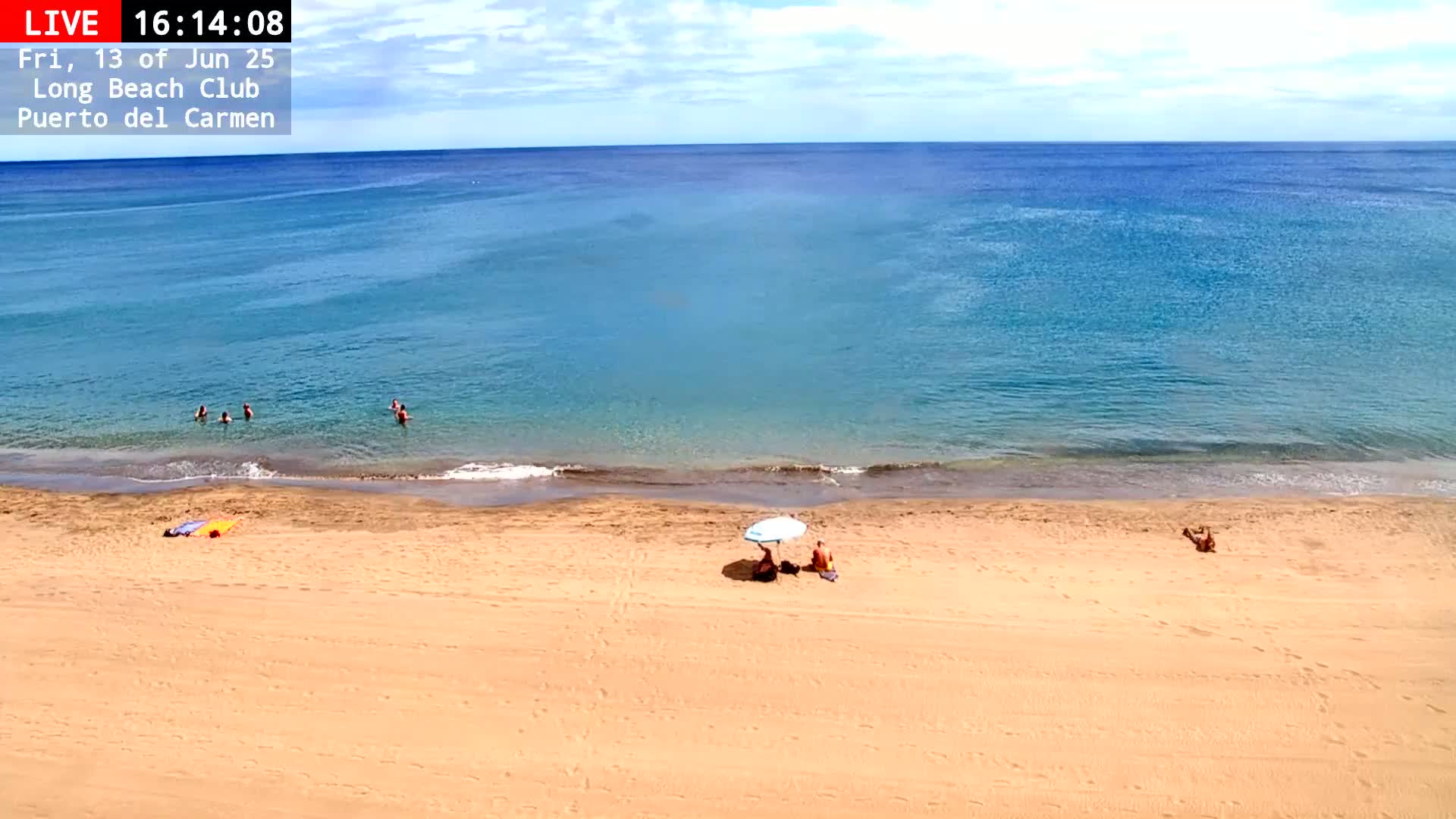 A sandy beach with a few people swimming in a calm, clear blue ocean under a mostly sunny sky.