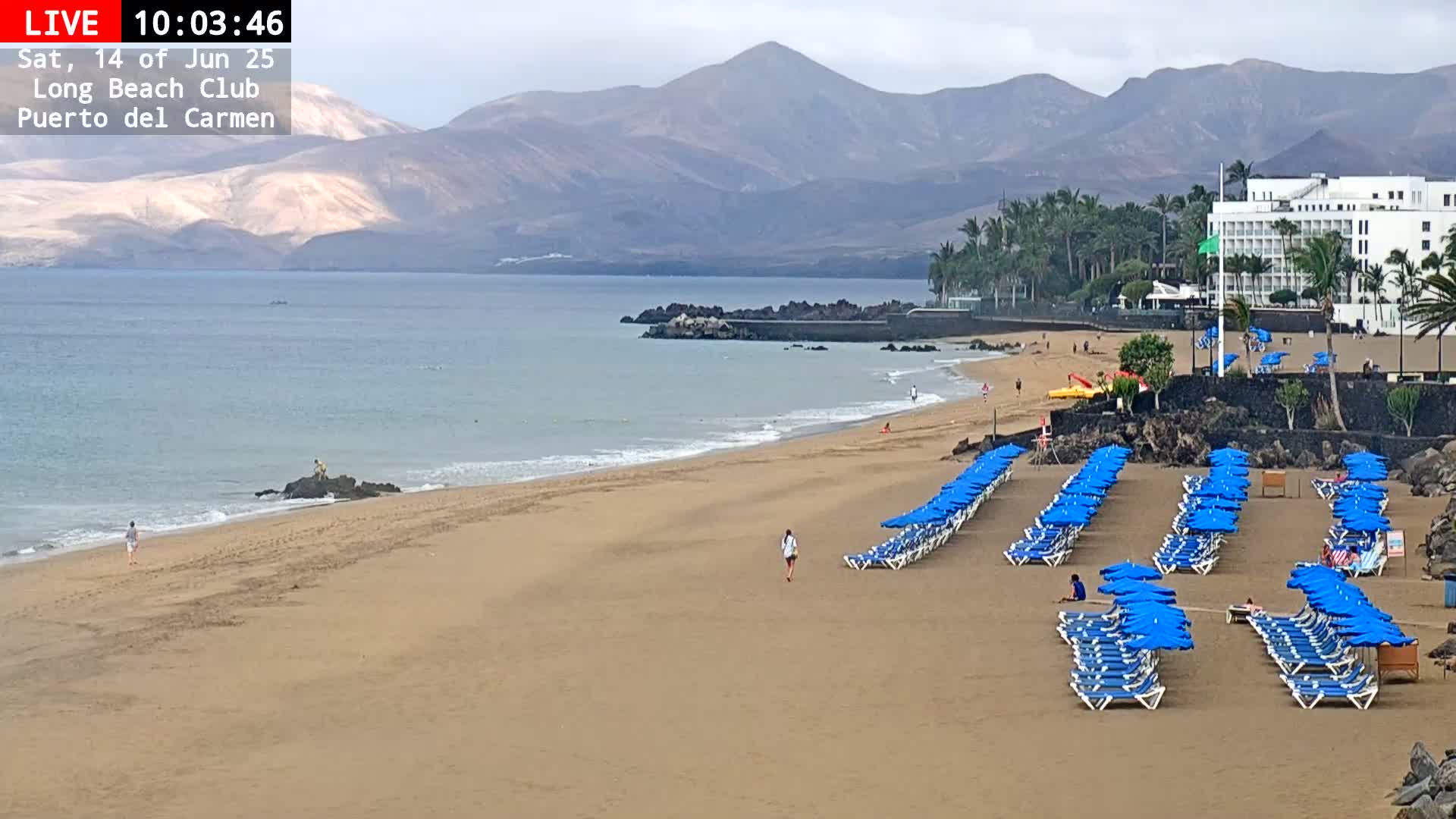 A sandy beach with rows of blue umbrellas and lounge chairs stretches along a calm ocean under an overcast sky, backed by a mountainous landscape and a large hotel.