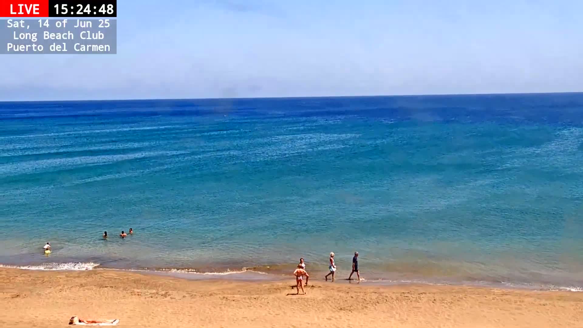 A sandy beach with a few people on it and in the calm, clear blue ocean under a sunny sky.