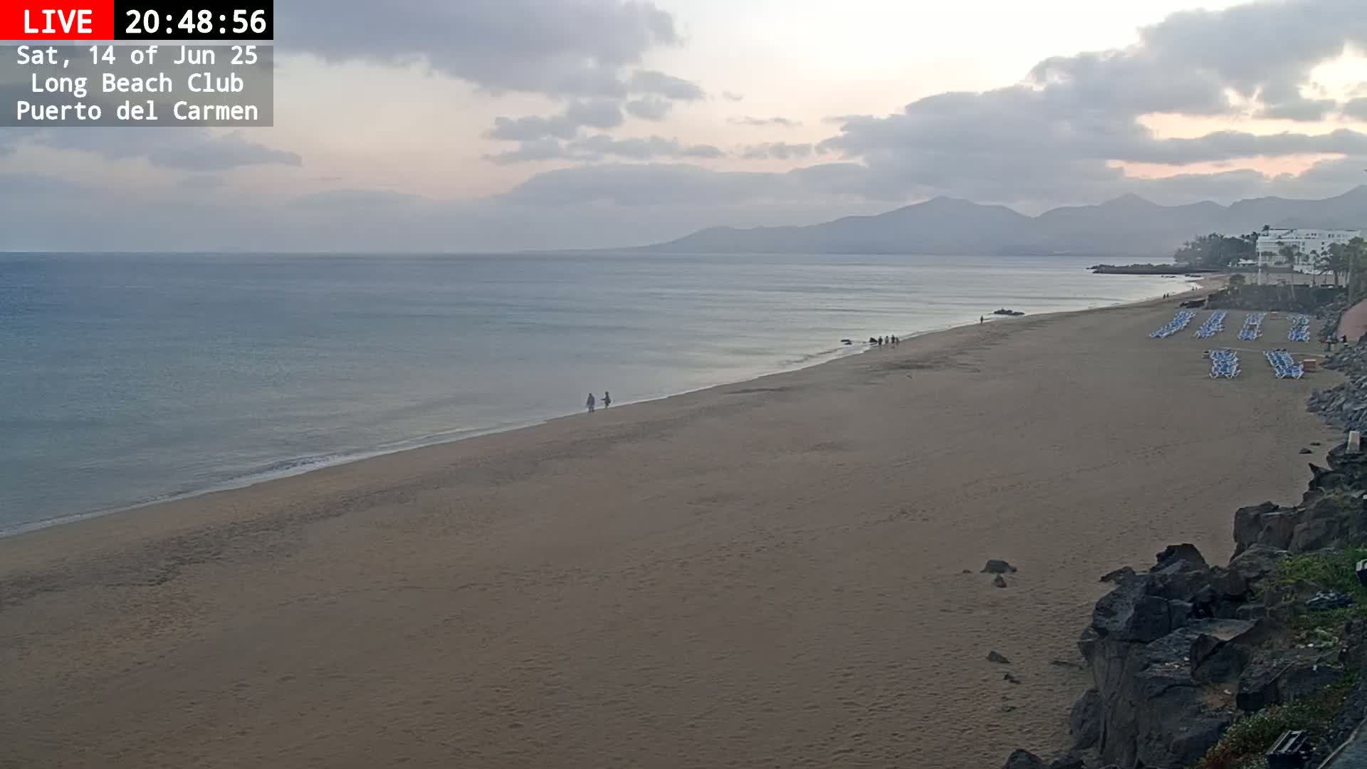 A sandy beach with a few people on it and in the calm, clear blue ocean under a sunny sky.