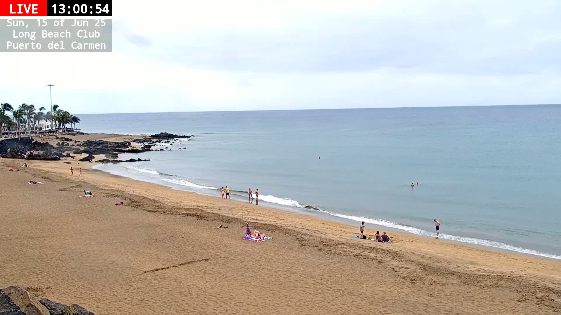 A mostly sunny day at a sandy beach with calm, light-blue water, palm trees, and a few people scattered on the sand and in the ocean.