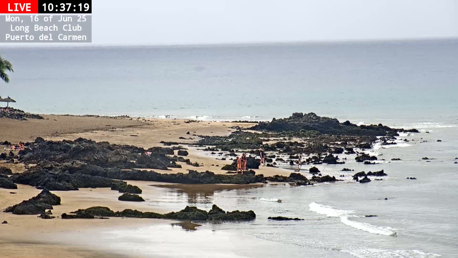 A sandy beach with dark rocks and a few people is visible, with calm, light-blue ocean waters under a hazy sky.