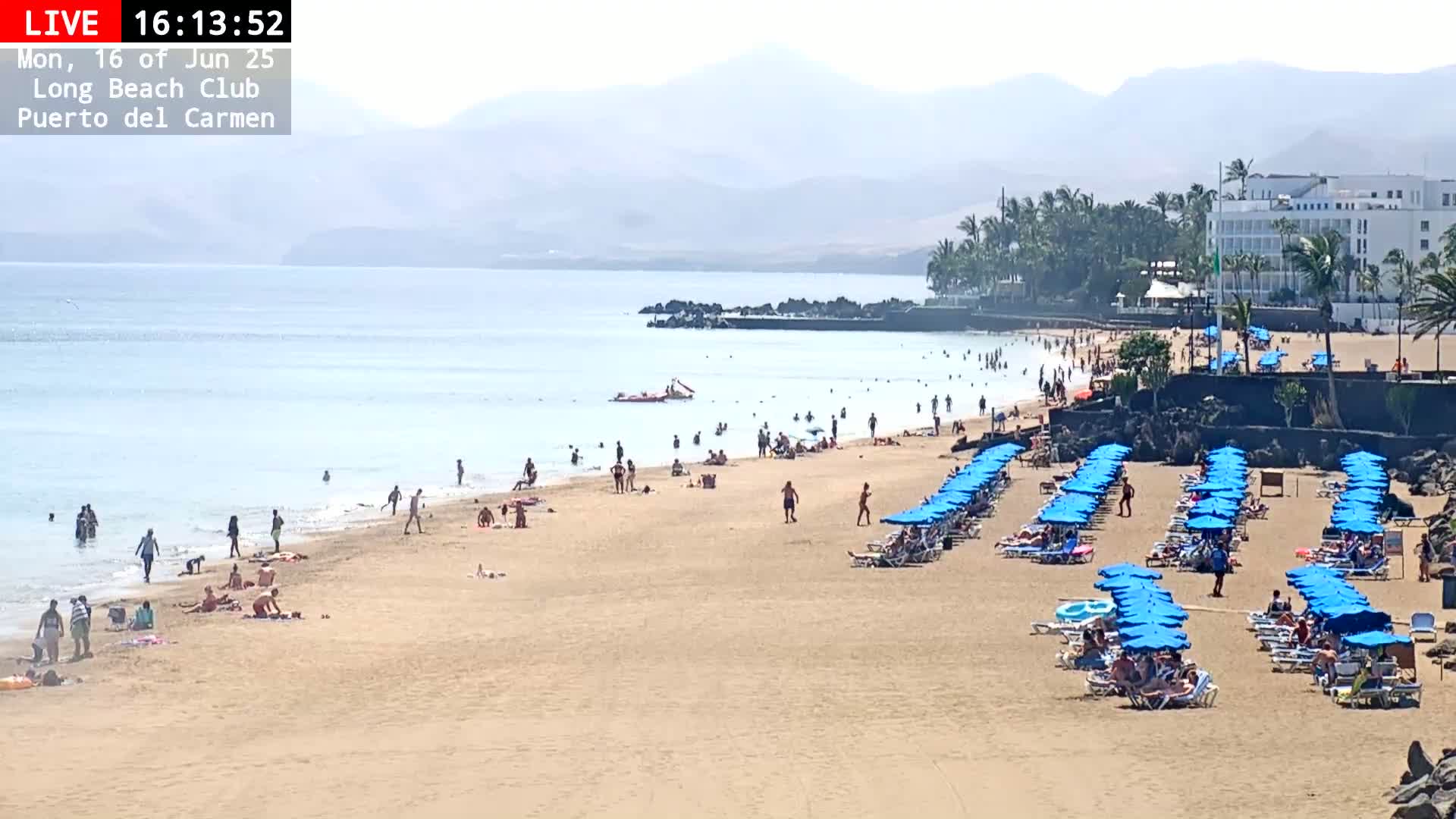 A sandy beach with many people swimming and sunbathing under numerous blue umbrellas, in front of a hotel and palm trees, on a hazy sunny day.