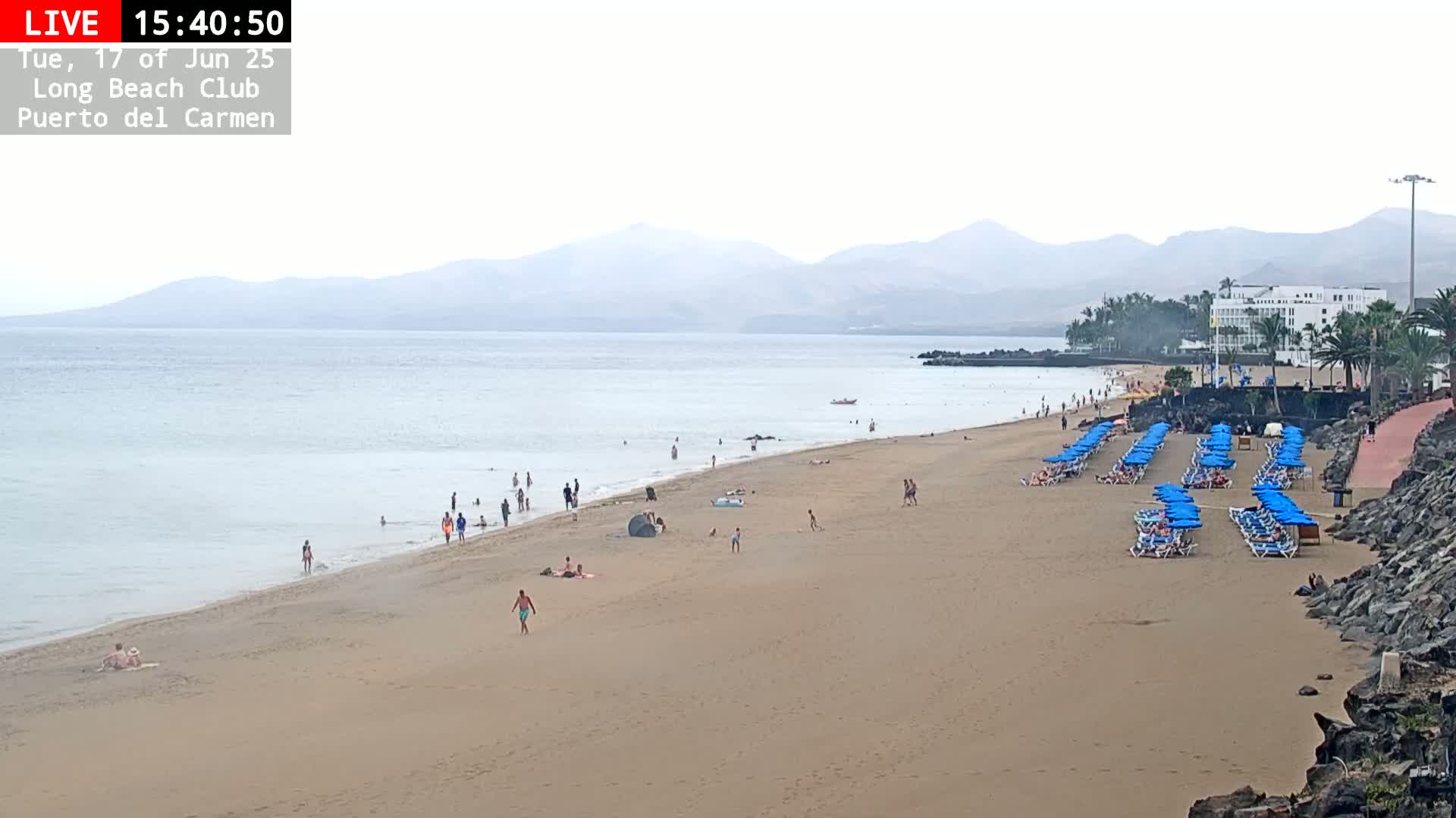 A sandy beach with scattered people, blue umbrellas and lounge chairs, and a calm ocean under an overcast sky, backed by distant mountains.