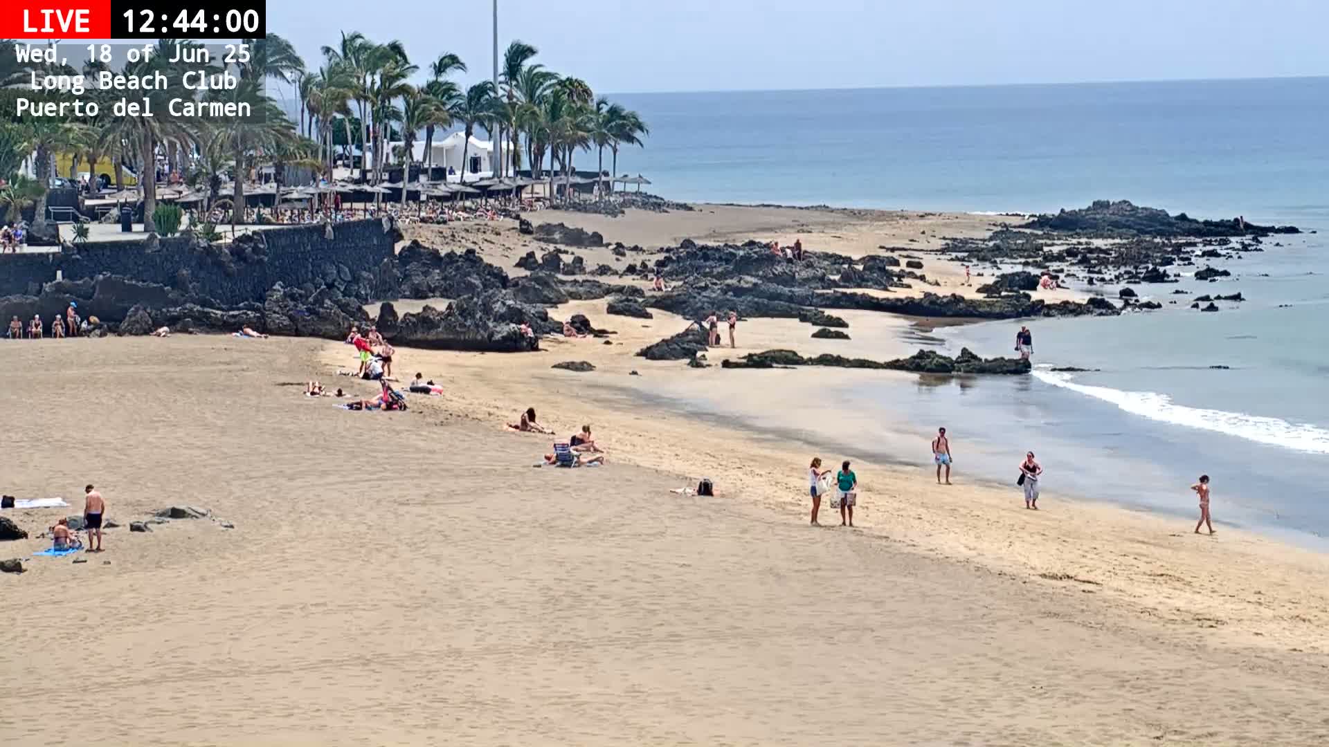 A sandy beach with a few people sunbathing and walking along the shore, under a sunny sky, is bordered by dark rocks and calm ocean waves.