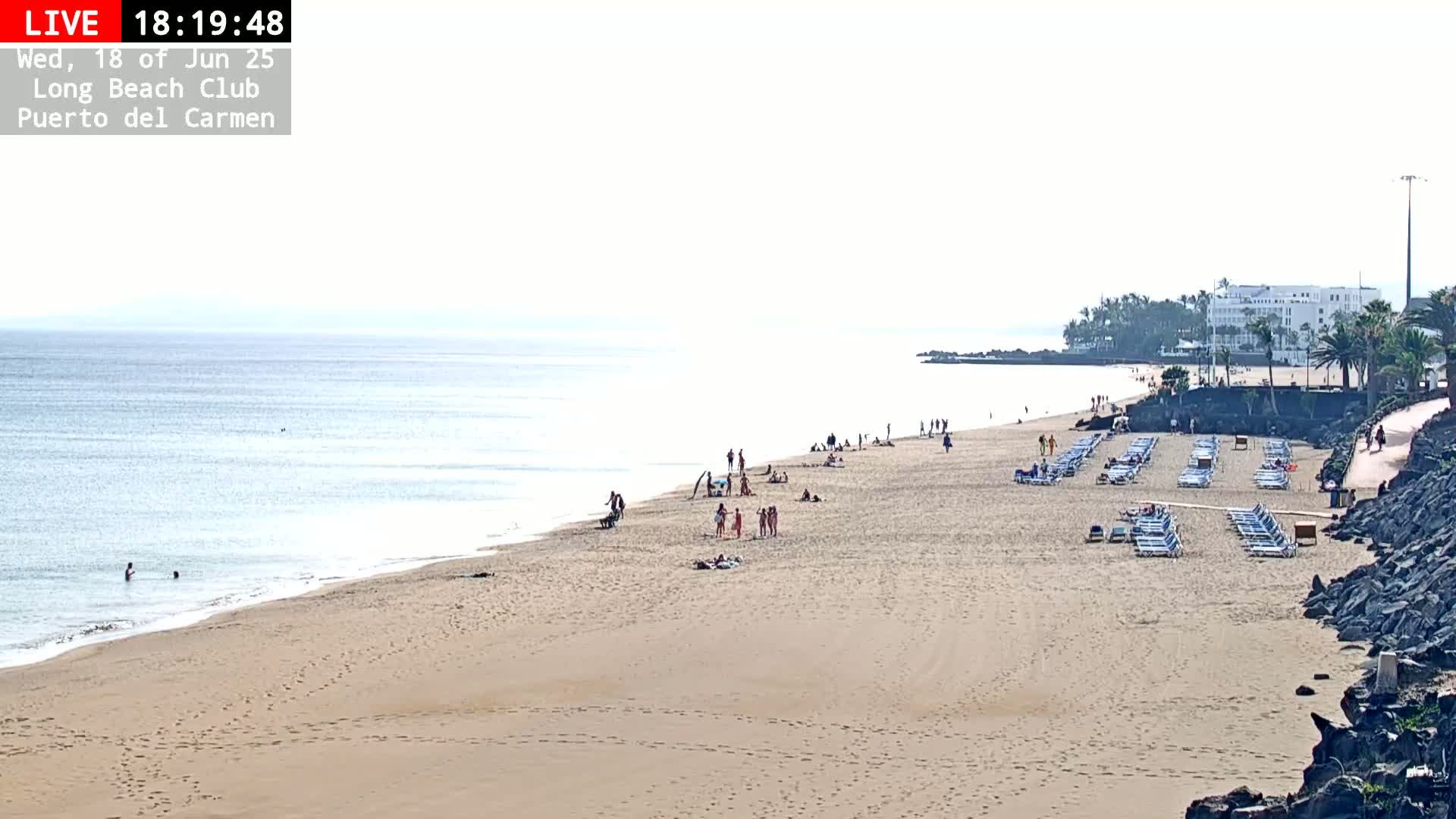 A sandy beach with a few people on it and rows of empty lounge chairs stretches along a calm ocean under a hazy sky.