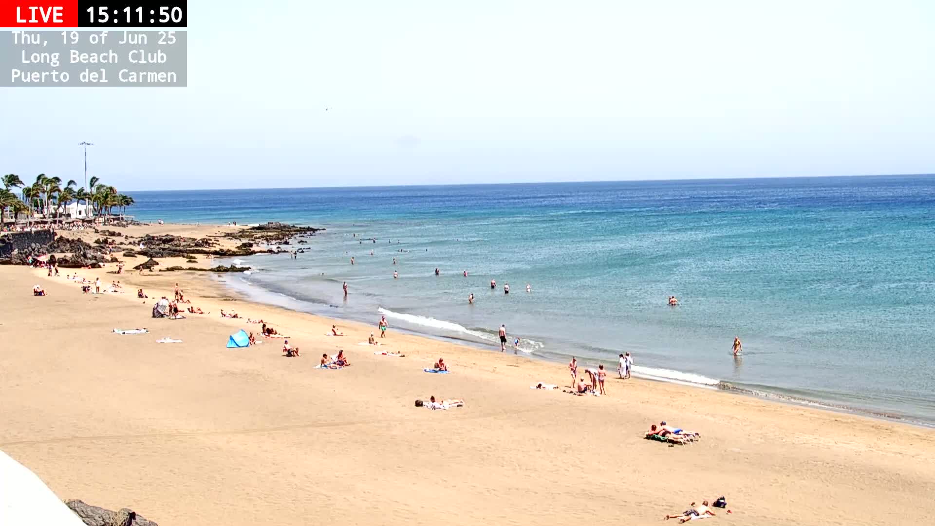 A sandy beach with many people sunbathing and swimming in a calm, turquoise ocean under a clear sky.