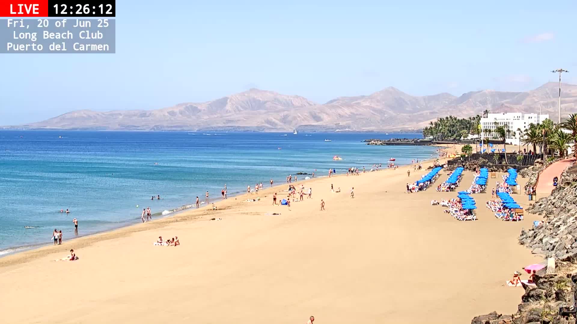 A sandy beach with many people sunbathing and swimming, lined with blue umbrellas and lounge chairs, stretches out to a calm turquoise ocean under a clear blue sky, backed by mountains.