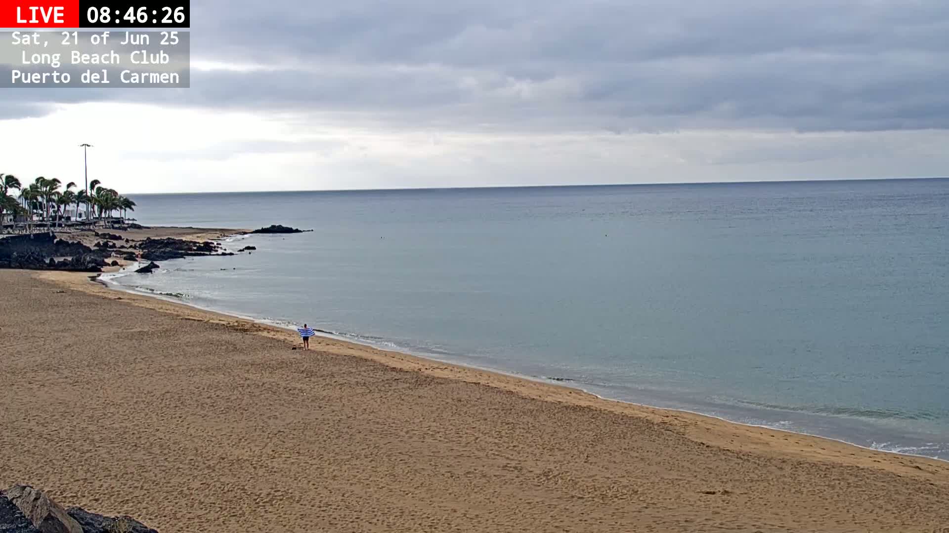 A person walks along a sandy beach next to a calm, light-blue ocean under an overcast sky.