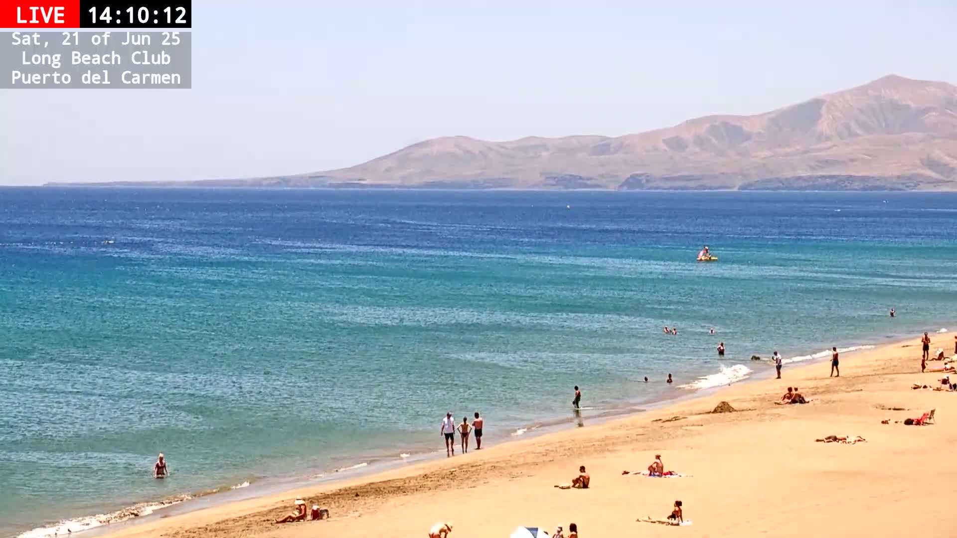 A sandy beach with people sunbathing and swimming in a calm, turquoise ocean under a clear sky, with mountains visible in the distance.