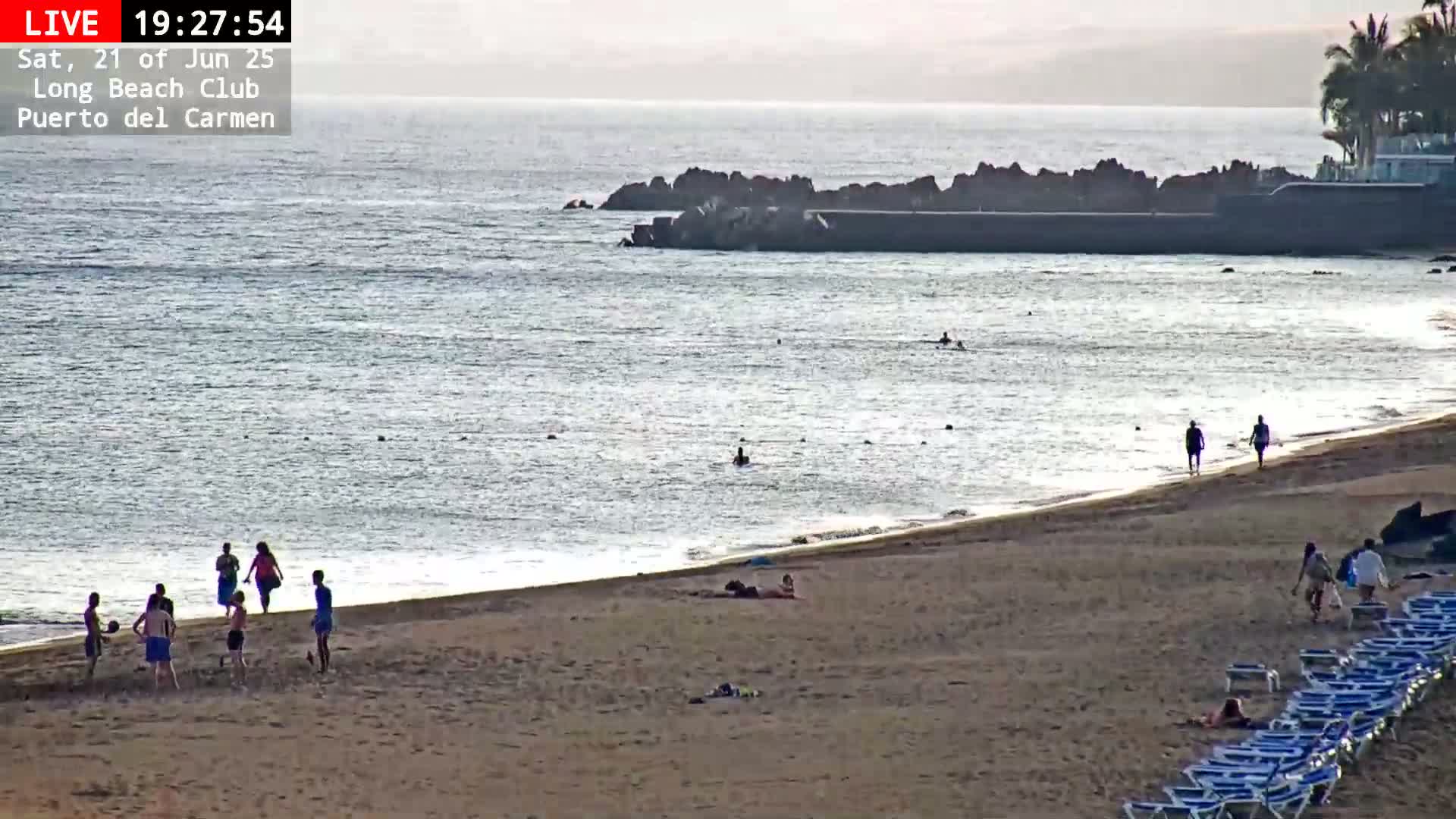 A sandy beach with people swimming in the ocean and others playing on the sand, under a mostly clear sky.