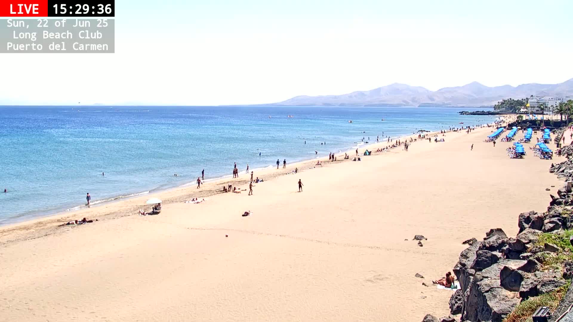 A sunny day at a sandy beach shows many people sunbathing and swimming in calm, blue water, with mountains visible in the distance.