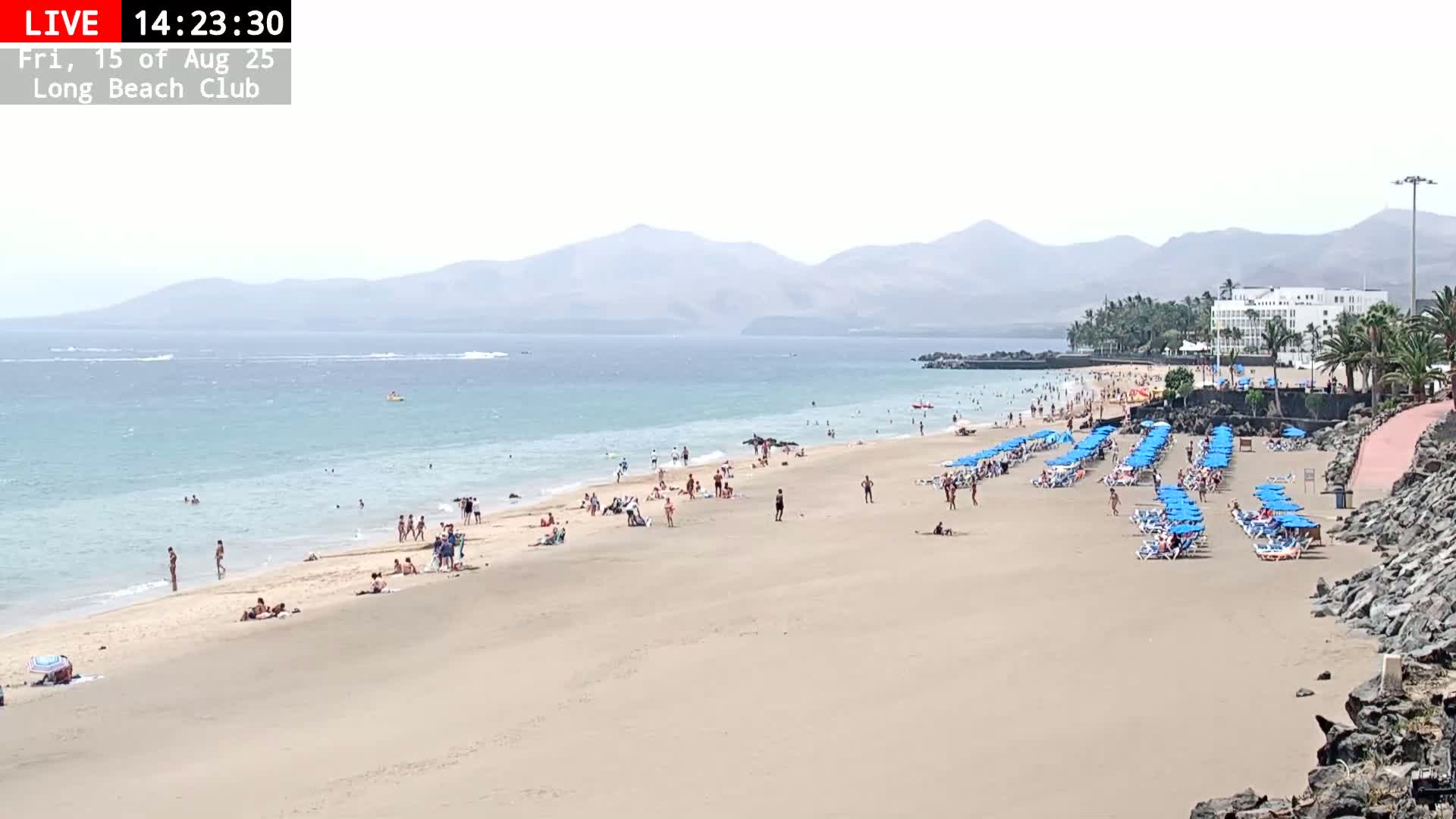 A sandy beach with a few people and rows of blue beach chairs stretches along a calm ocean under a mostly cloudy sky, mountains visible in the distance.