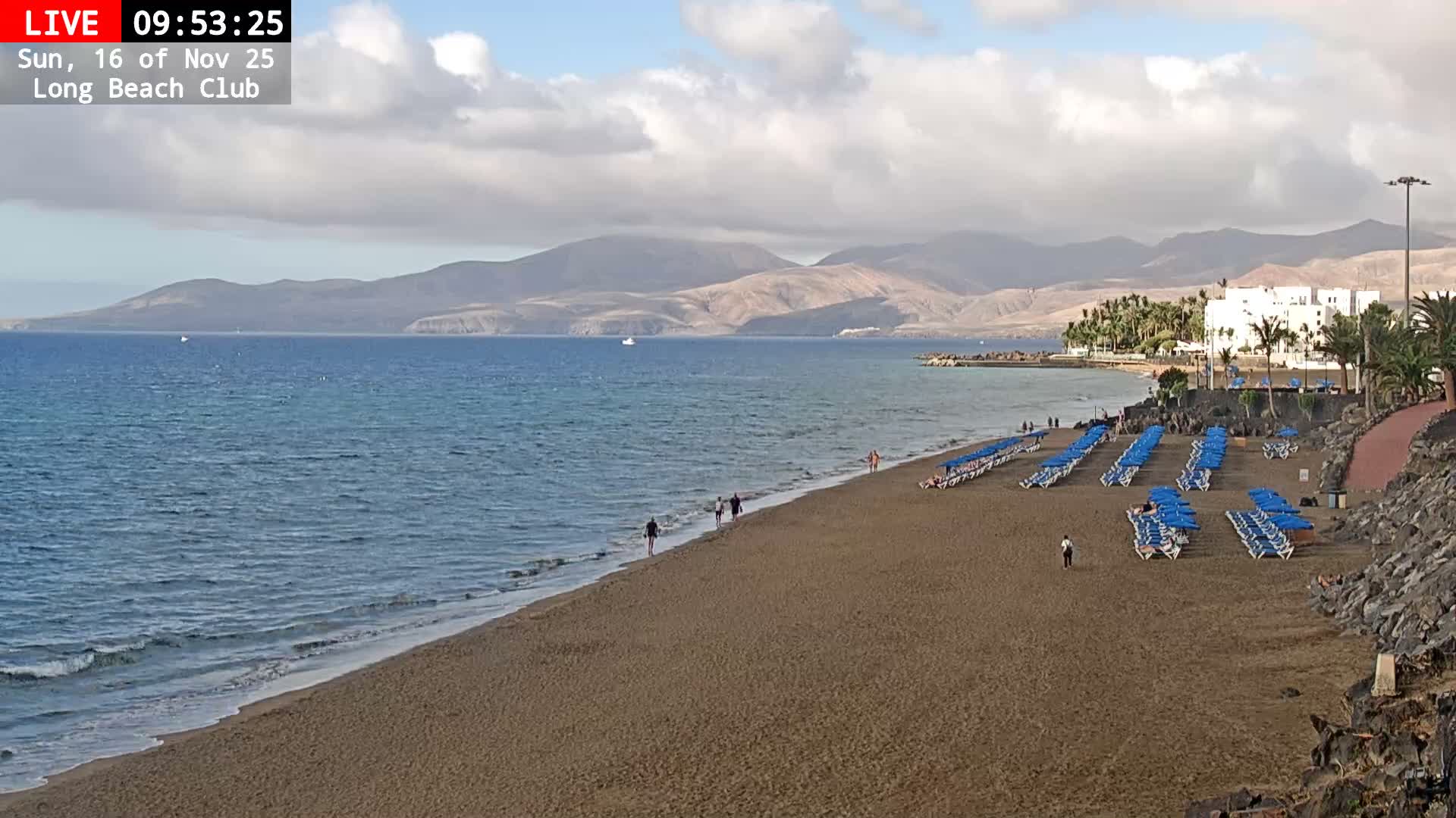 A scenic view captures a wide sandy beach lined with numerous blue sun loungers and a few people strolling by the calm ocean, with distant mountains and coastal buildings visible under a partly cloudy sky.