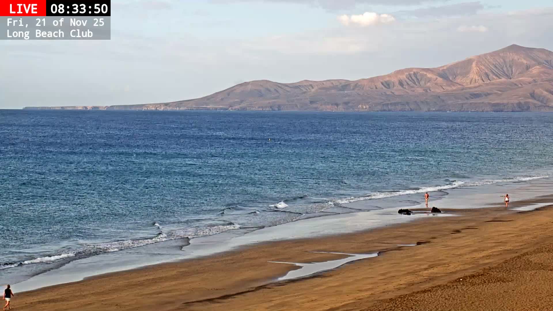 A sandy beach with gentle waves is seen under a partly cloudy sky, with several people strolling near the waterline and arid mountains in the distant background across the blue sea.
