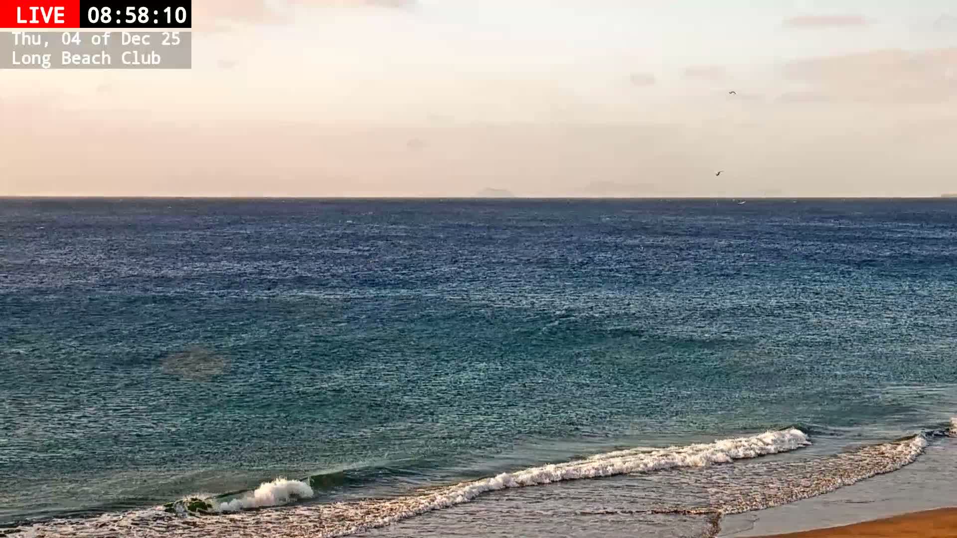 Calm blue-green ocean waters with gentle white-capped waves roll towards the bottom of the frame under a clear, warm-toned, and slightly hazy sky, with faint land visible on the distant horizon.
