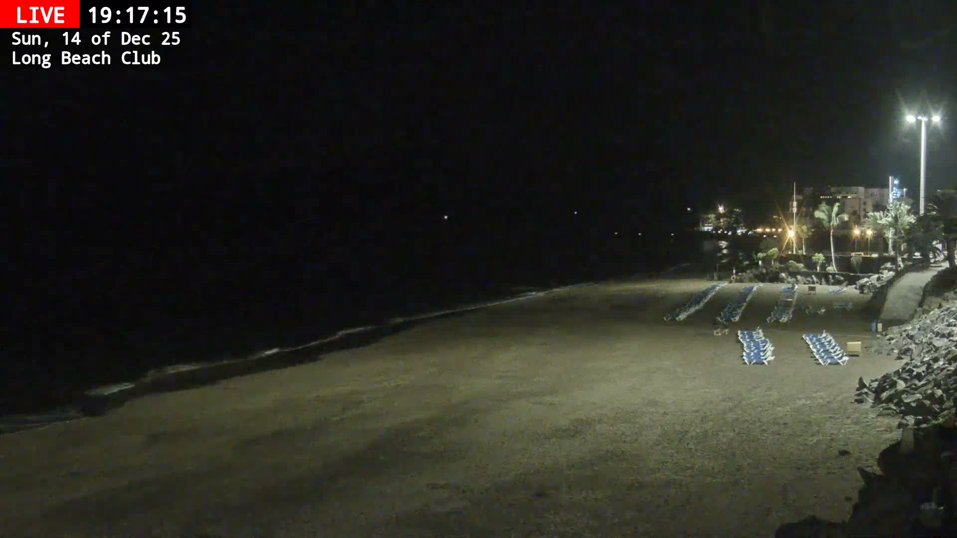 A bustling beach with people swimming and relaxing on the sand, backed by palm trees and rocky formations, under a clear, sunny sky.