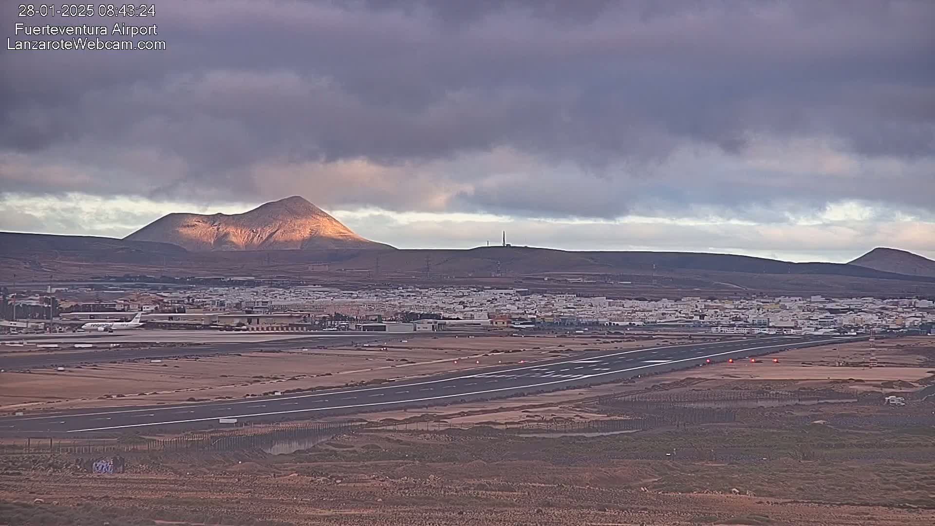 An airport runway and surrounding buildings are seen under a cloudy sky, with volcanic mountains in the background.