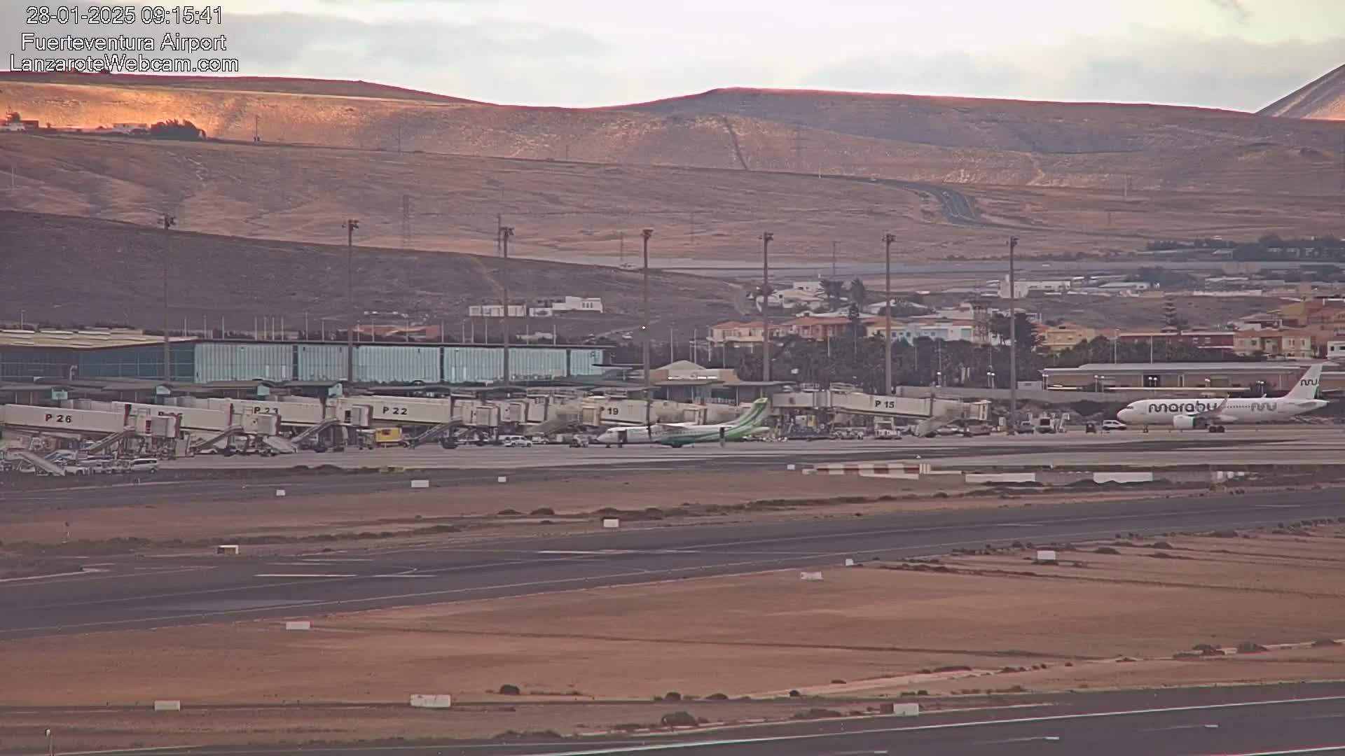 An airport tarmac with several airplanes and jet bridges, situated against a backdrop of arid, tan-colored hills under a mostly clear sky.