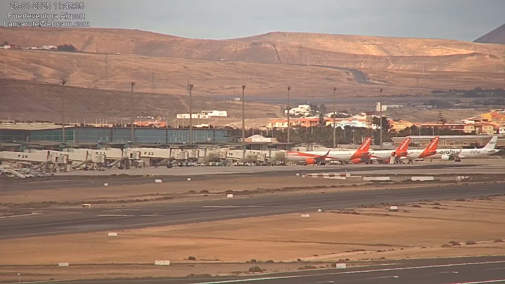Several airplanes are parked at gates at an airport under an overcast sky, with a dry, hilly landscape visible in the background.