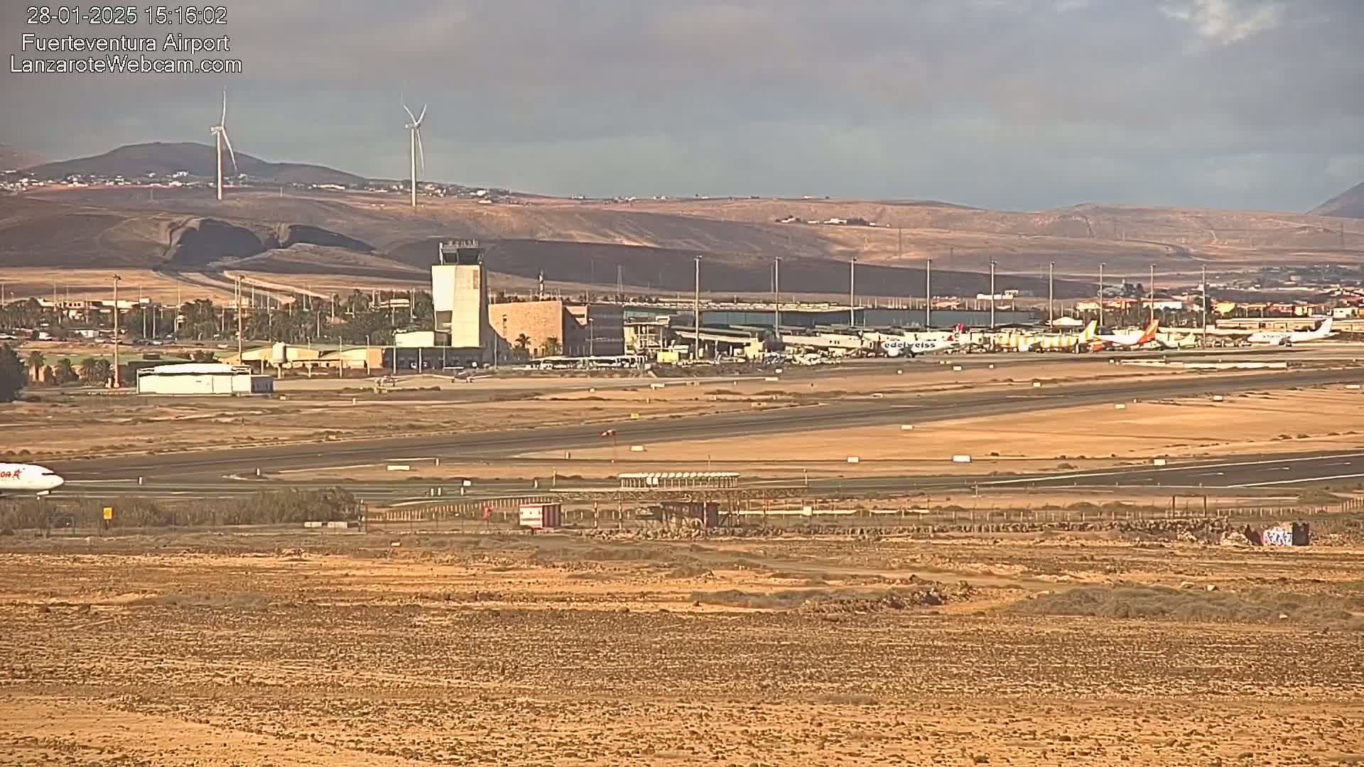 A long shot of an airport under a partly cloudy sky, showing planes on the tarmac, airport buildings, and wind turbines in the distance.