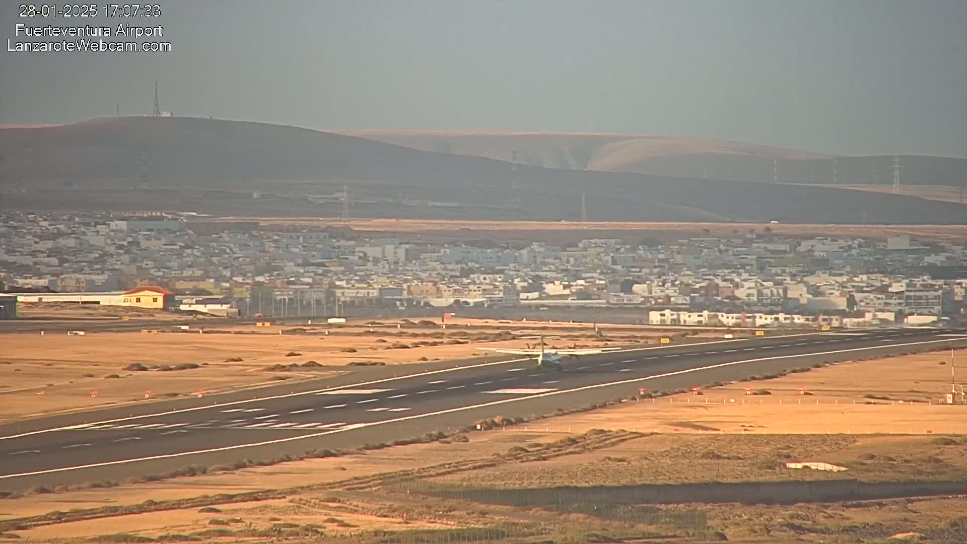 A small airplane taxis on a runway at an airport near a town, under hazy, sunny conditions.