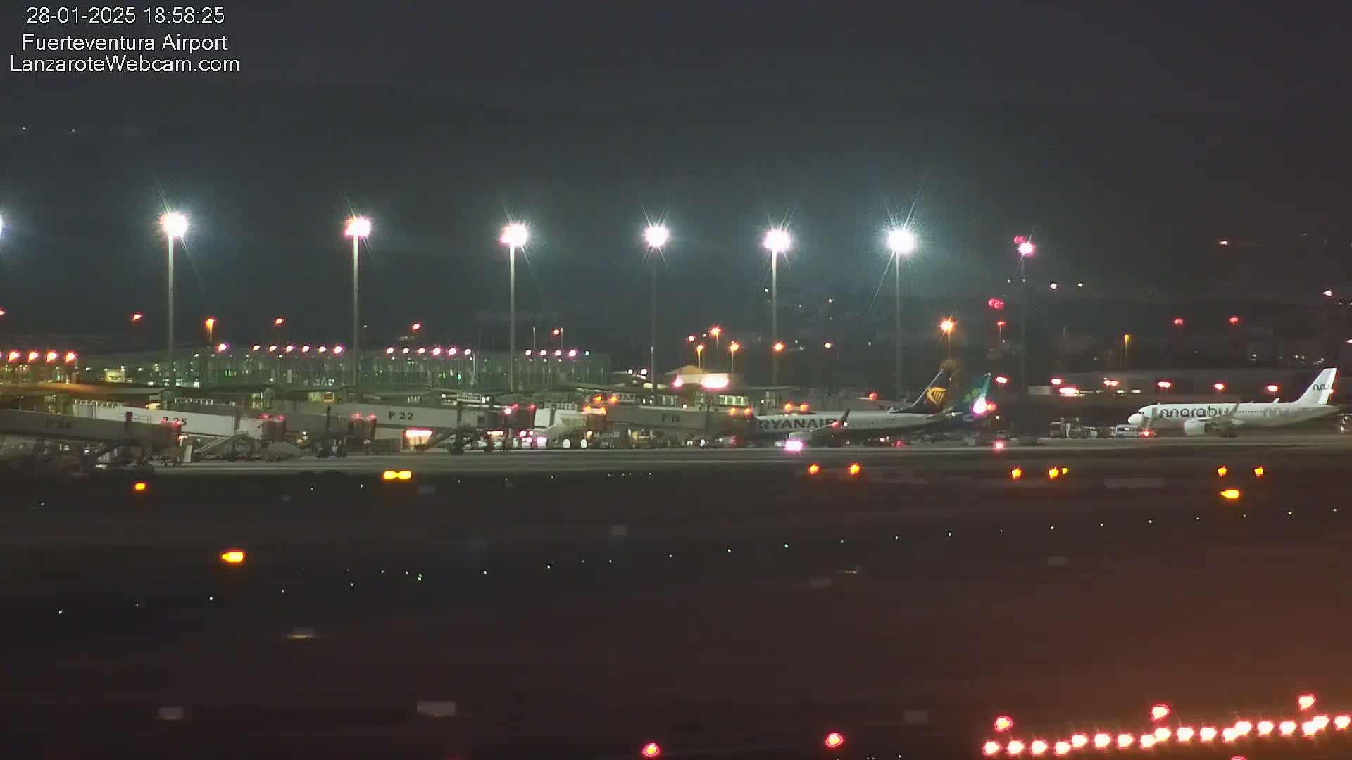 At night, under clear skies, two airplanes are parked at a brightly lit airport terminal with several jet bridges connected.