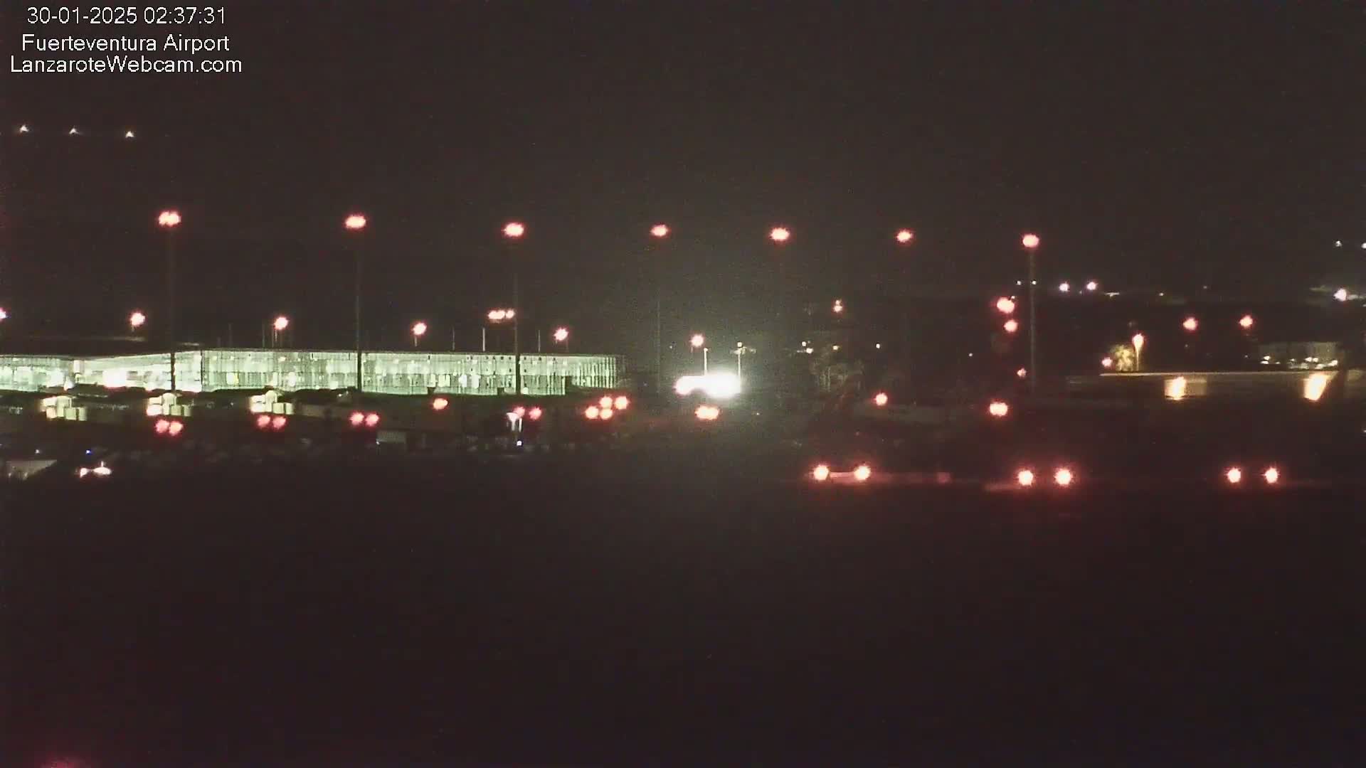 At night, a long, brightly lit building is visible beyond a road with several vehicles, all under a dark sky.