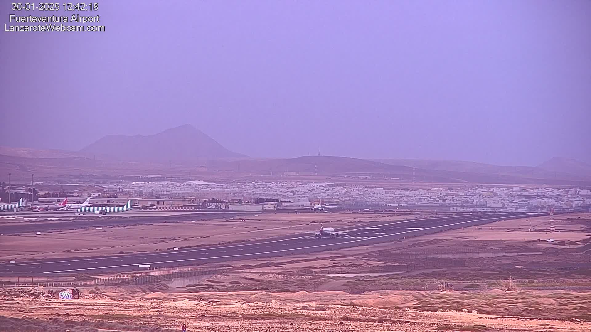 An airport runway with several airplanes, a nearby town, and hazy mountains in the distance under a hazy, purplish sky.
