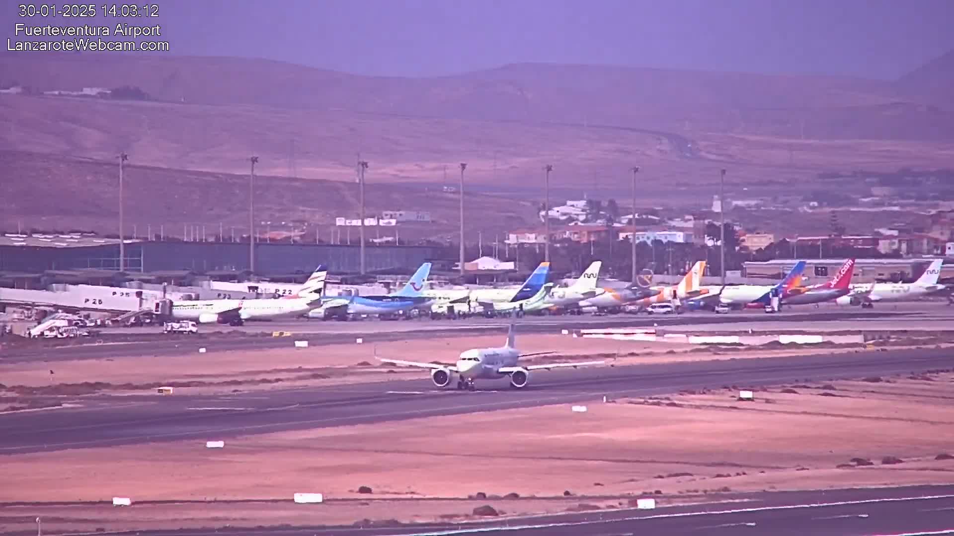 A single airplane taxis on a runway past numerous other parked airplanes at an airport under hazy, overcast conditions.