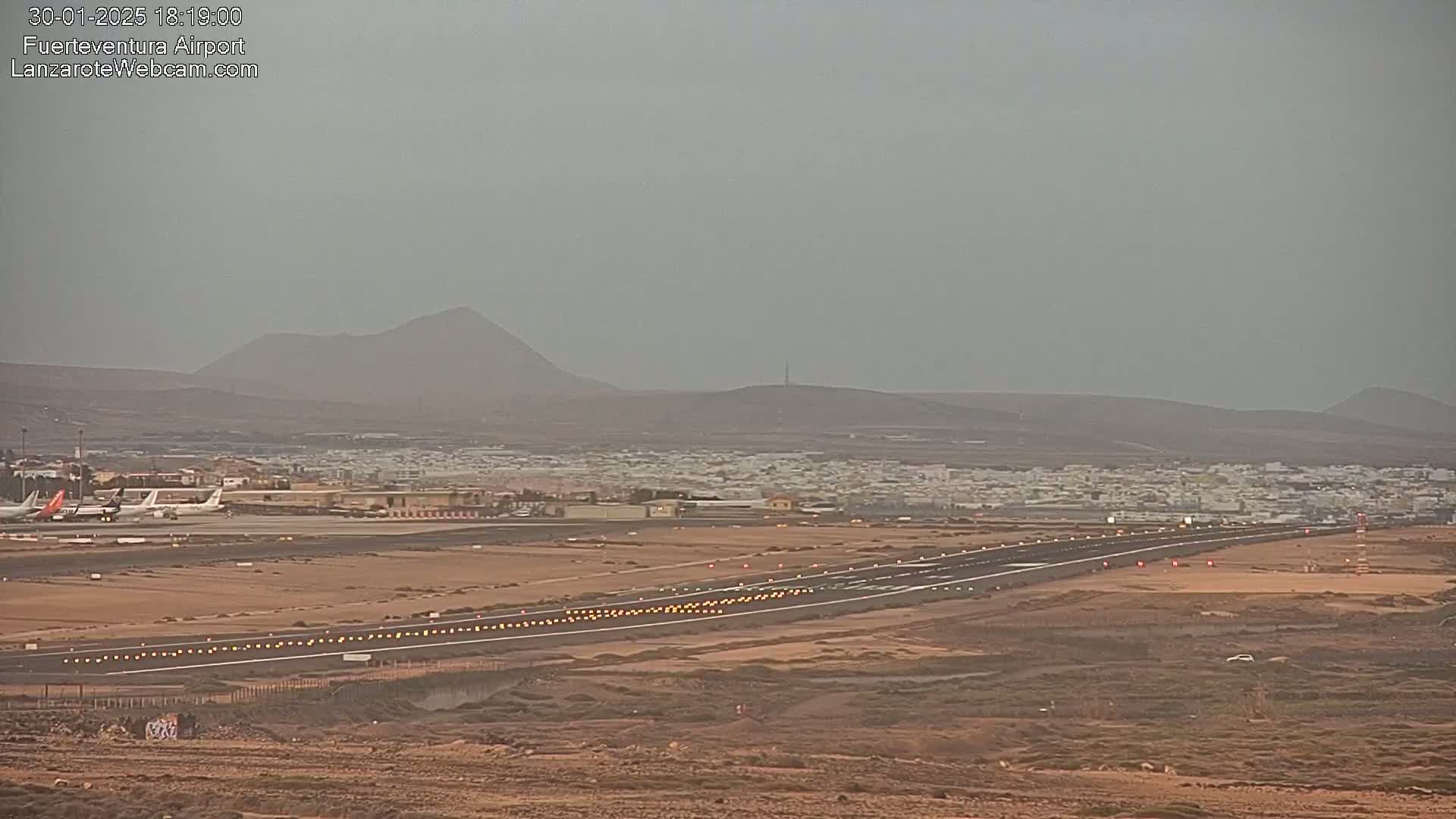 A long, flat runway at an airport, with several airplanes parked nearby, is seen under hazy skies with distant mountains in the background.
