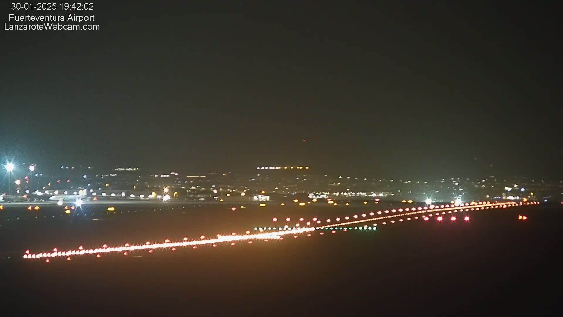 A nighttime view of an airport runway illuminated with lights, showing aircraft lights and the surrounding area faintly lit in the distance under a dark sky.