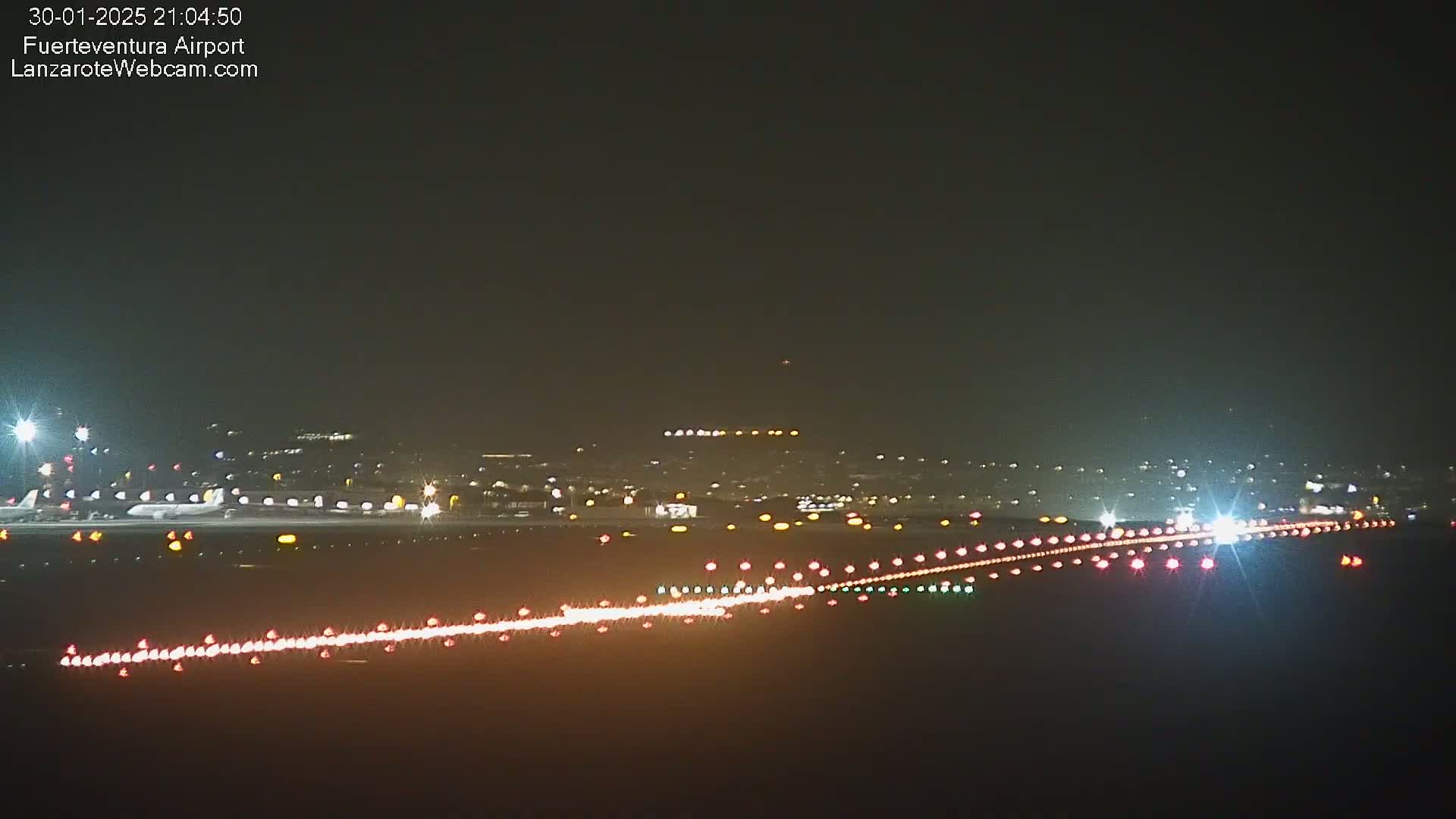 A nighttime long shot of an airport runway with several planes on the tarmac and lights illuminating the runway.