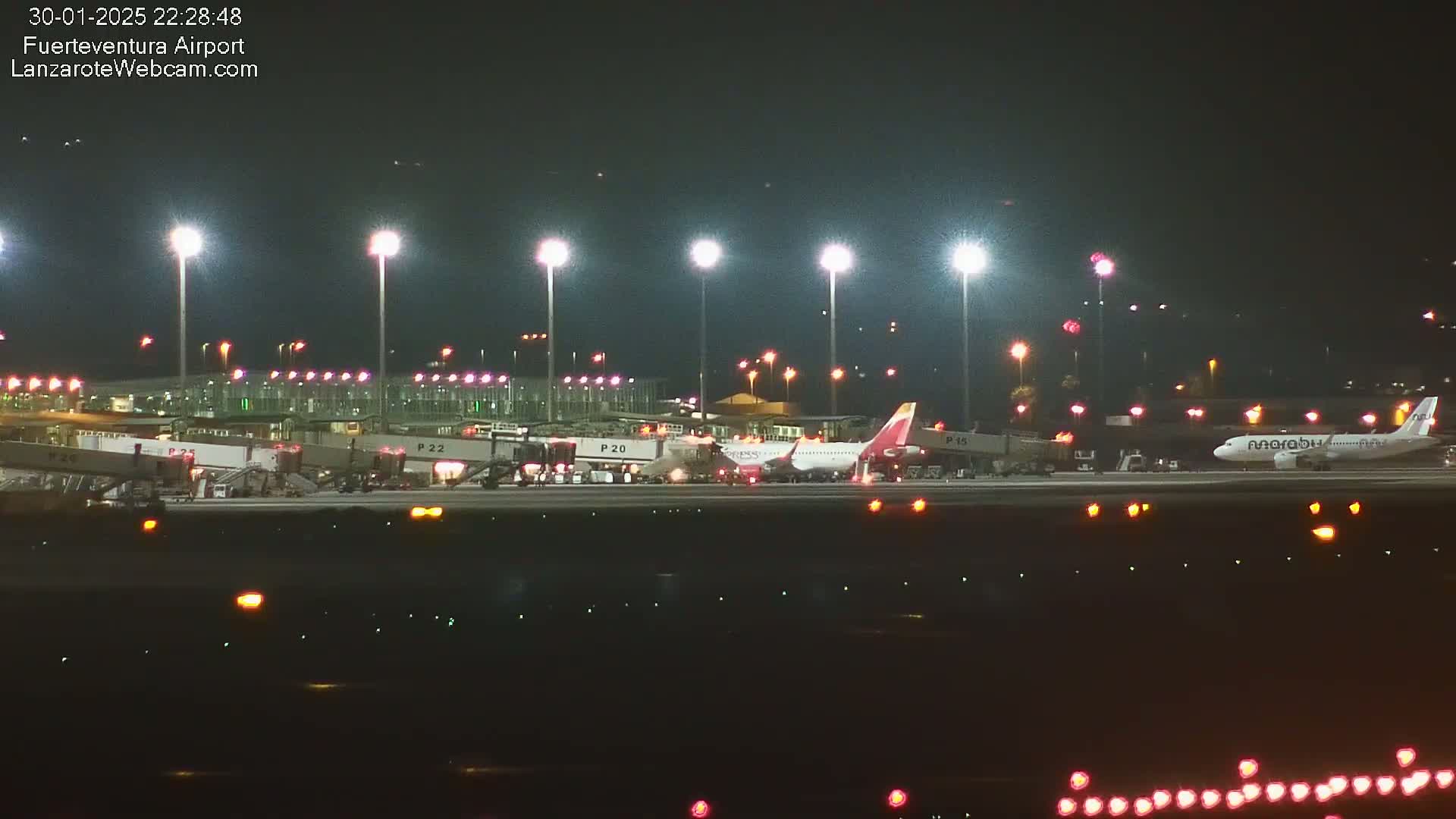 At night, two airplanes are parked at an airport gate under bright lights, with several jet bridges visible.