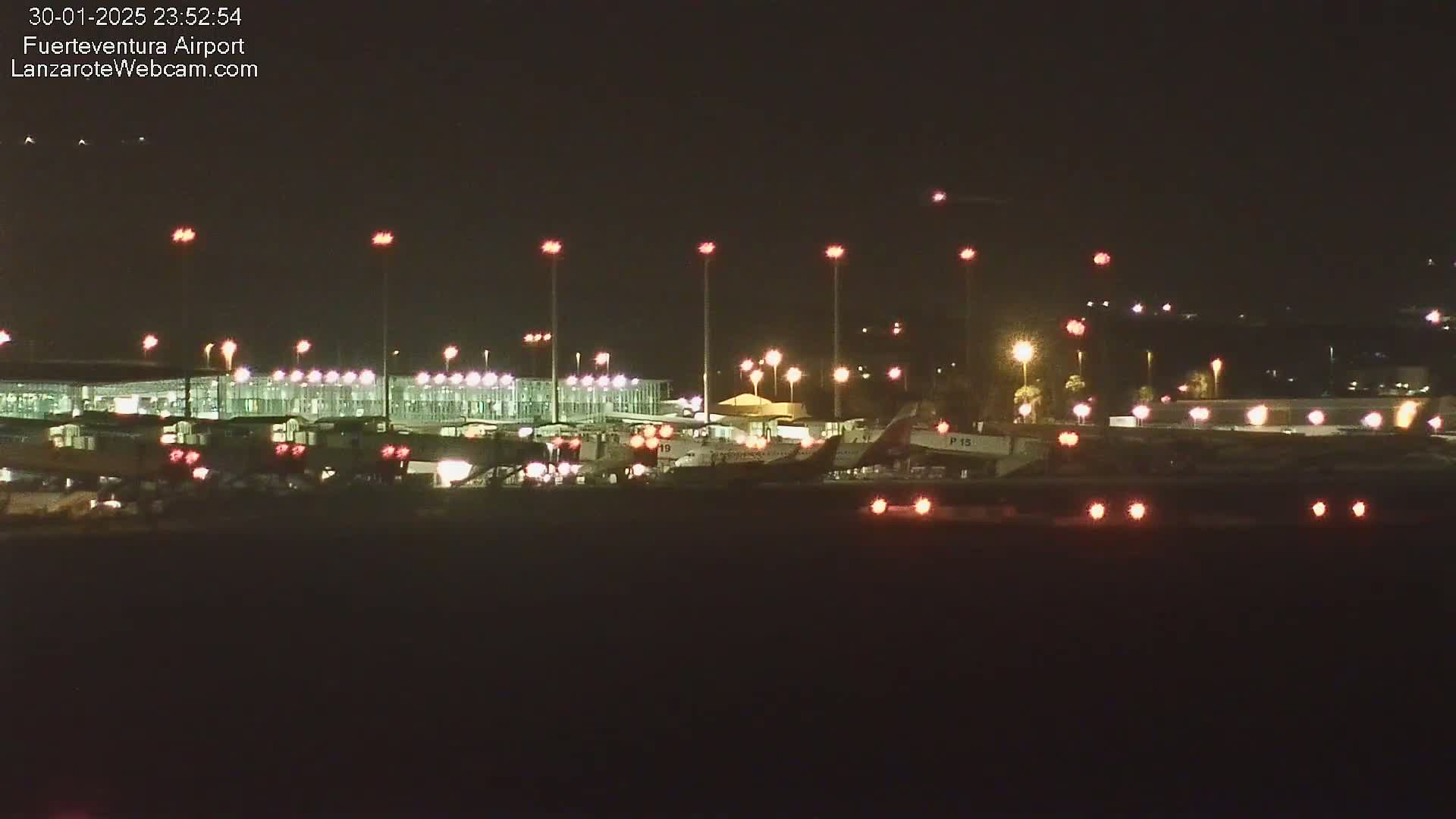 At night, a brightly lit airport terminal and several airplanes are visible on the tarmac under a clear, dark sky.