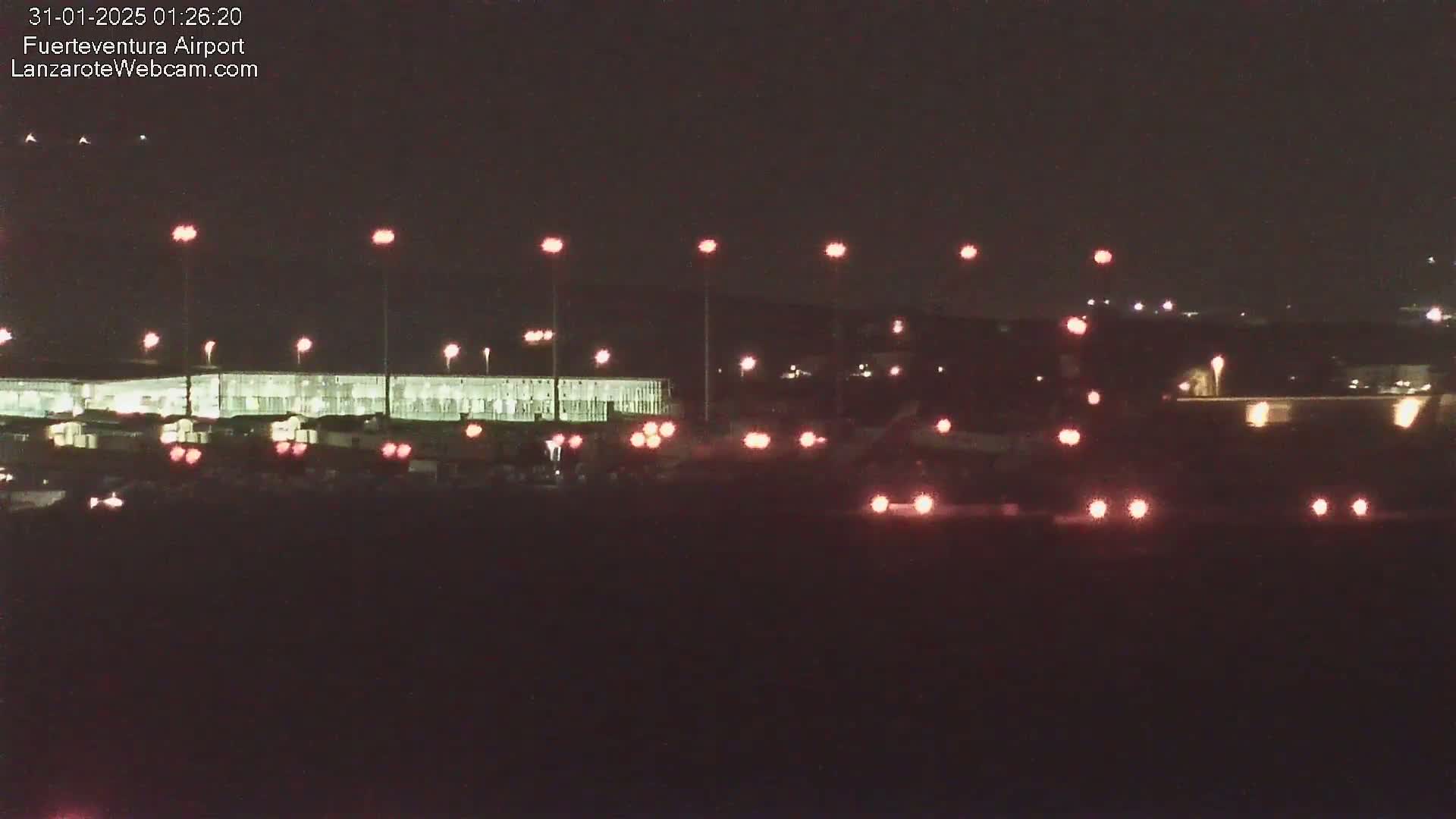 A nighttime view of an airport terminal building illuminated with lights, and various vehicles with their taillights visible on the tarmac.