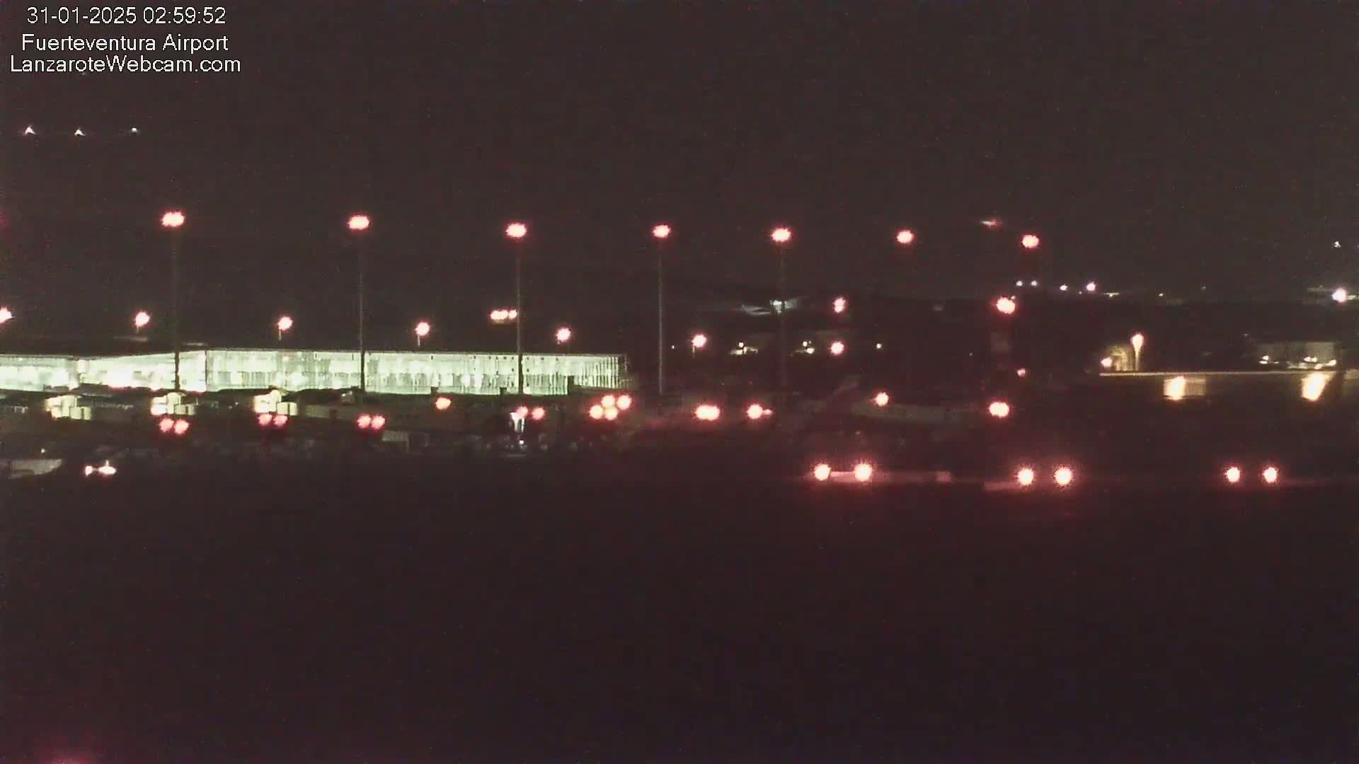 A nighttime view of an airport tarmac with numerous lights illuminating vehicles and buildings under a dark sky.