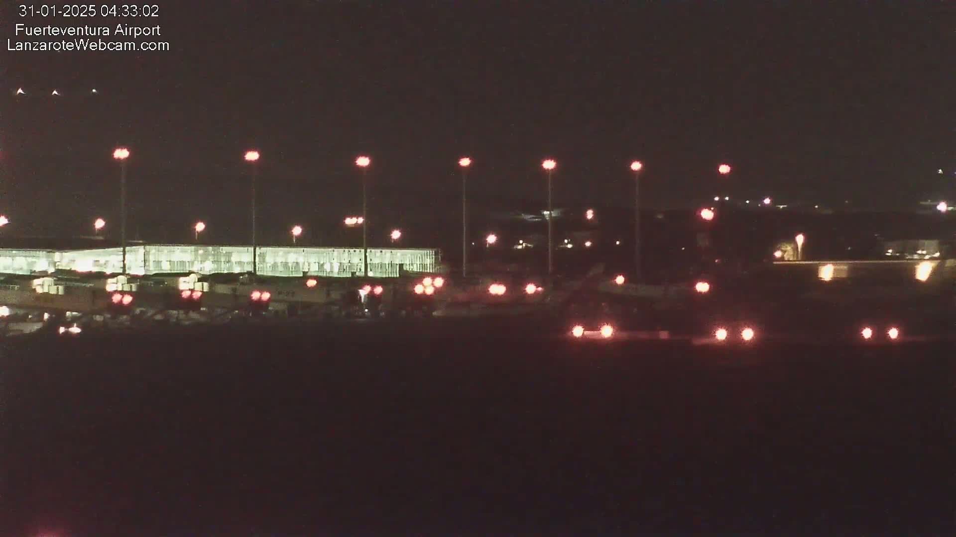 A nighttime view of an airport terminal building illuminated by lights, with vehicles visible in the foreground under a dark sky.